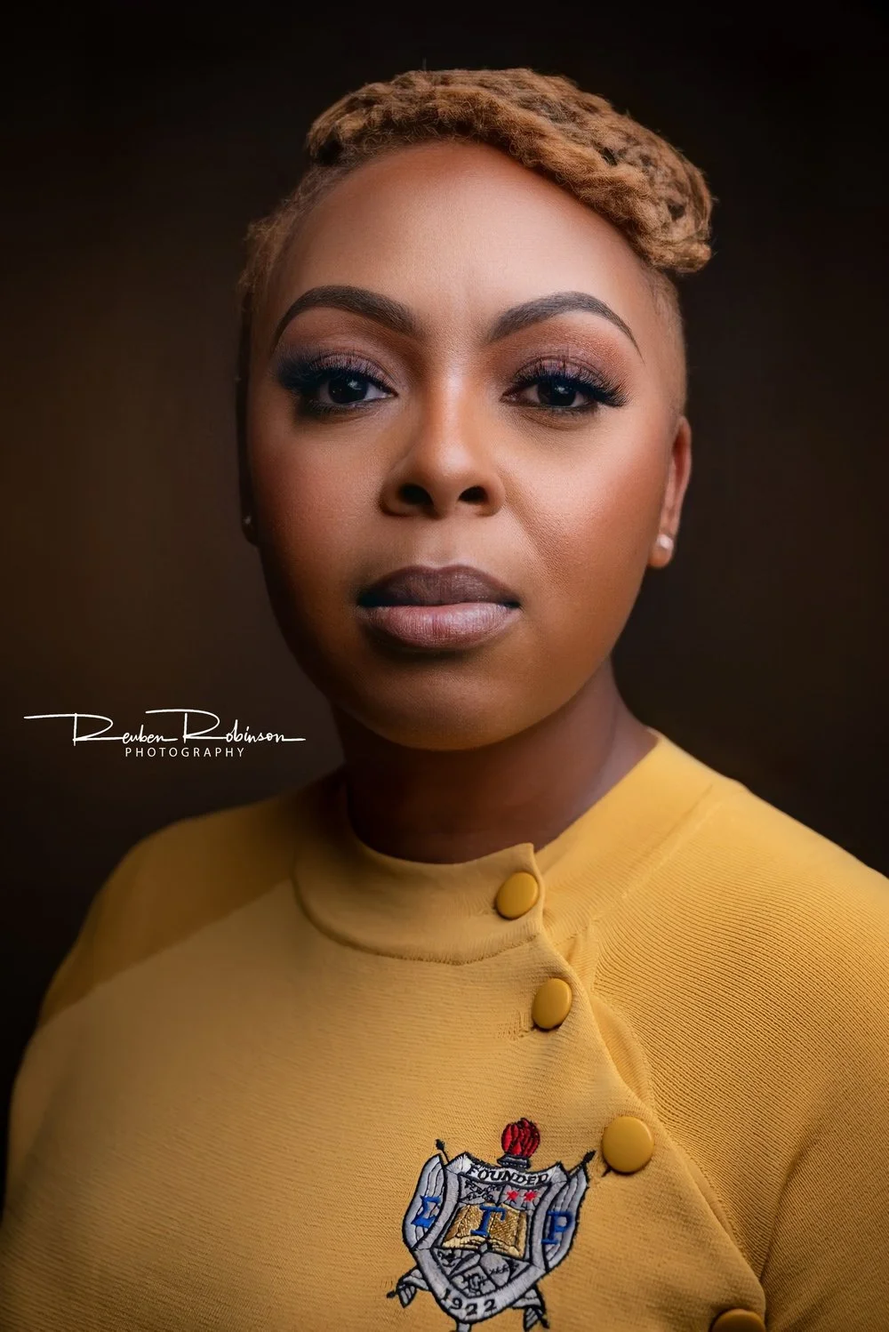 A portrait of a woman with short, styled hair, wearing a yellow top with decorative buttons and an embroidered emblem on the chest, against a dark background.