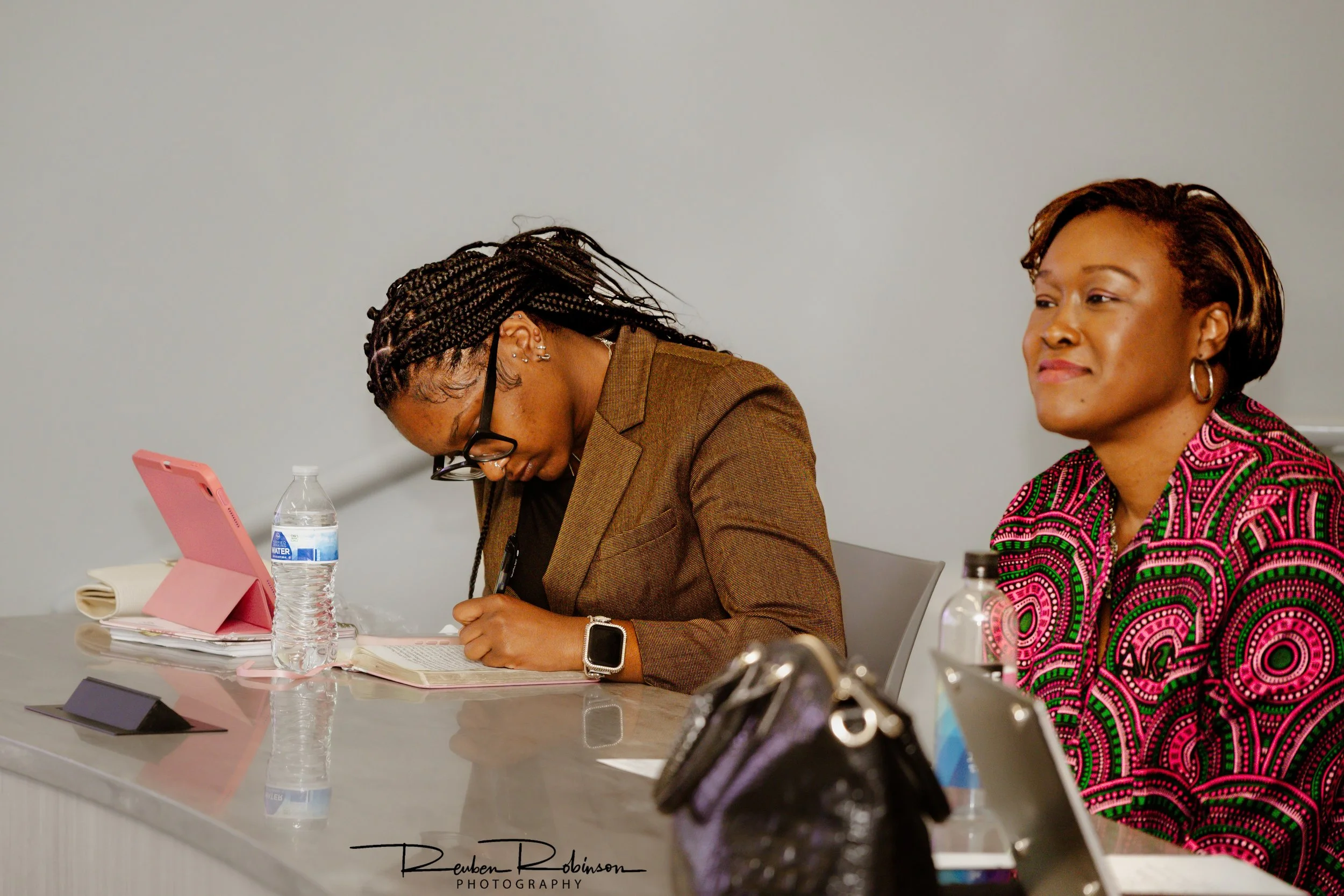 Two women sitting at a conference table, one wearing glasses and a brown blazer, the other wearing a colorful patterned dress, with water bottles, notebooks, and tablets on the table.