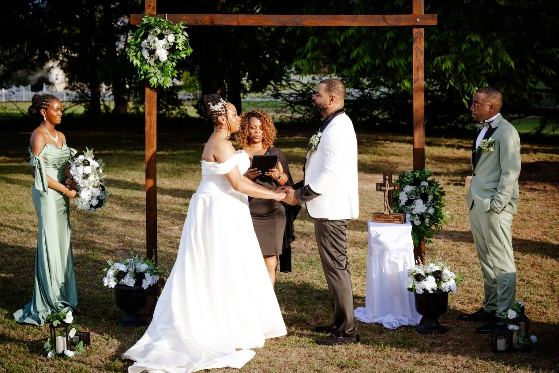 A couple getting married outdoors under a wooden arch decorated with flowers, with officiant, bridesmaid, and groomsman present, surrounded by greenery.