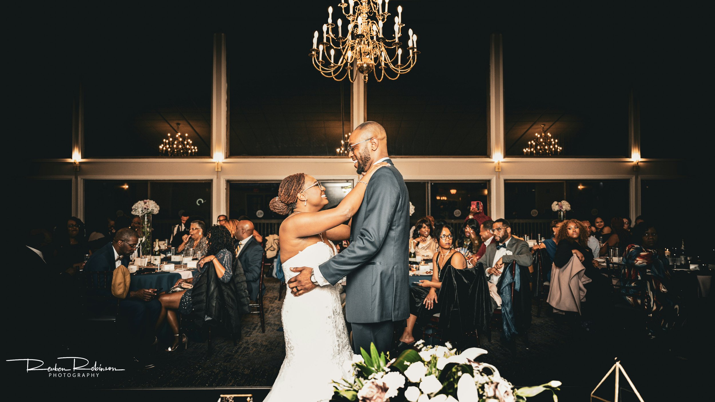 A newlywed couple dancing at their wedding reception, surrounded by seated guests. The venue is elegantly decorated with chandeliers and floral arrangements in the foreground.