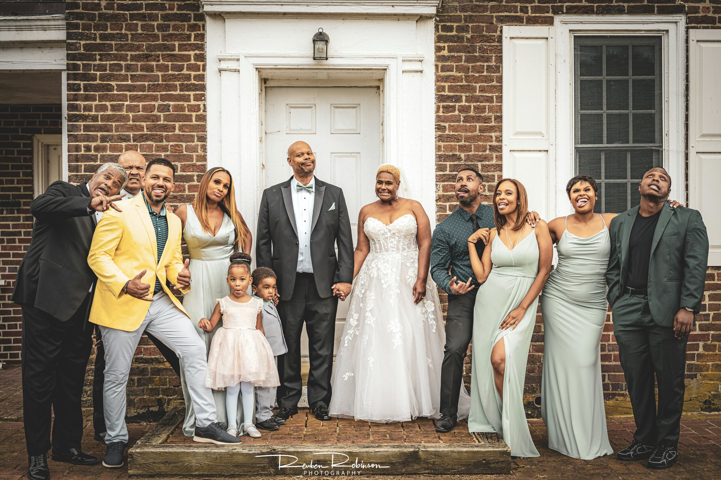Group photo of a diverse wedding party in front of a brick building. The bride is in a white dress, and the groom is in a black suit. Attendees include adults and two children, with some dressed in formal and colorful attire, making playful expressio