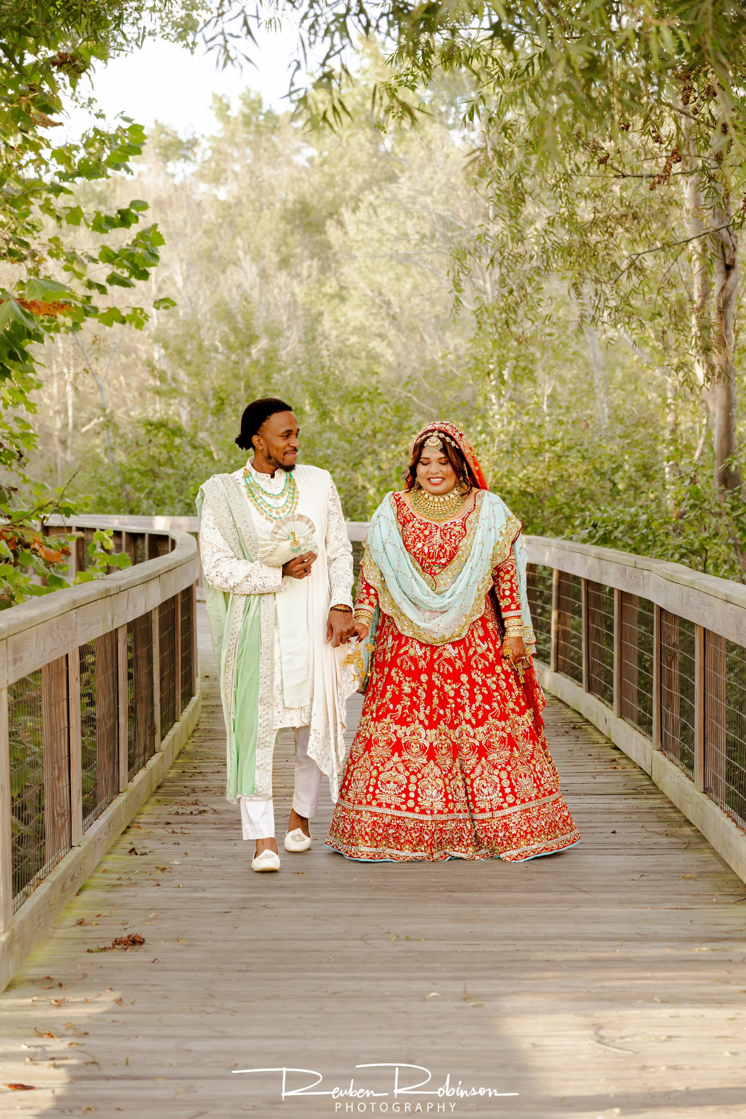 Bride and groom walking on a wooden bridge surrounded by green trees, dressed in traditional South Asian wedding attire.