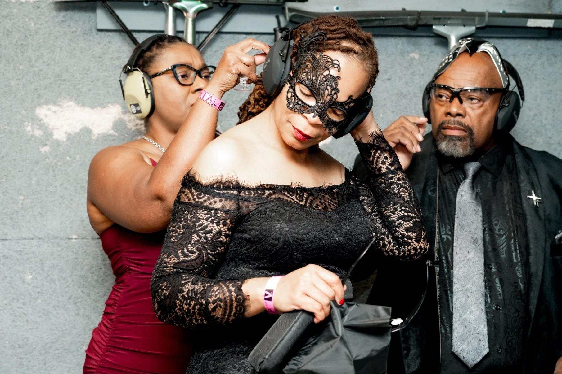 Three people wearing headphones, with one woman in an intricate black lace mask and dress adjusting her headphones, standing in front of a textured gray wall.