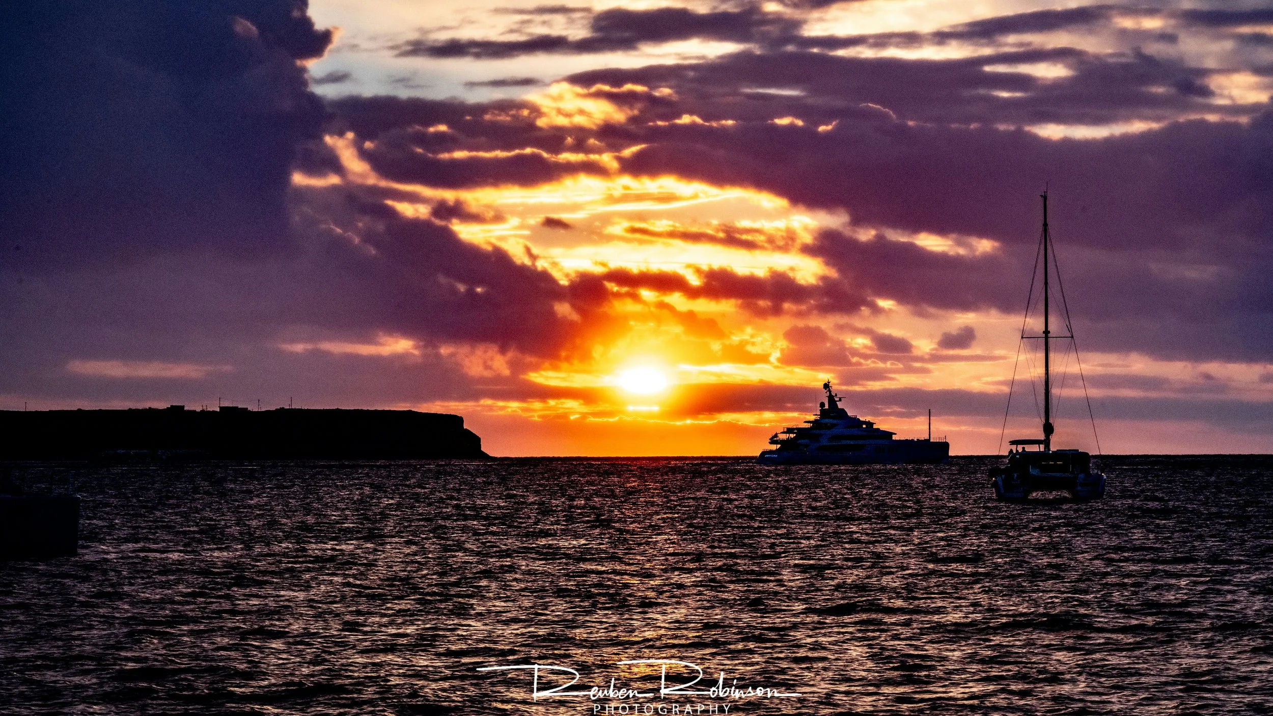 Sunset over the water with two boats, one sailboat and one yacht, silhouetted against a sky filled with clouds.