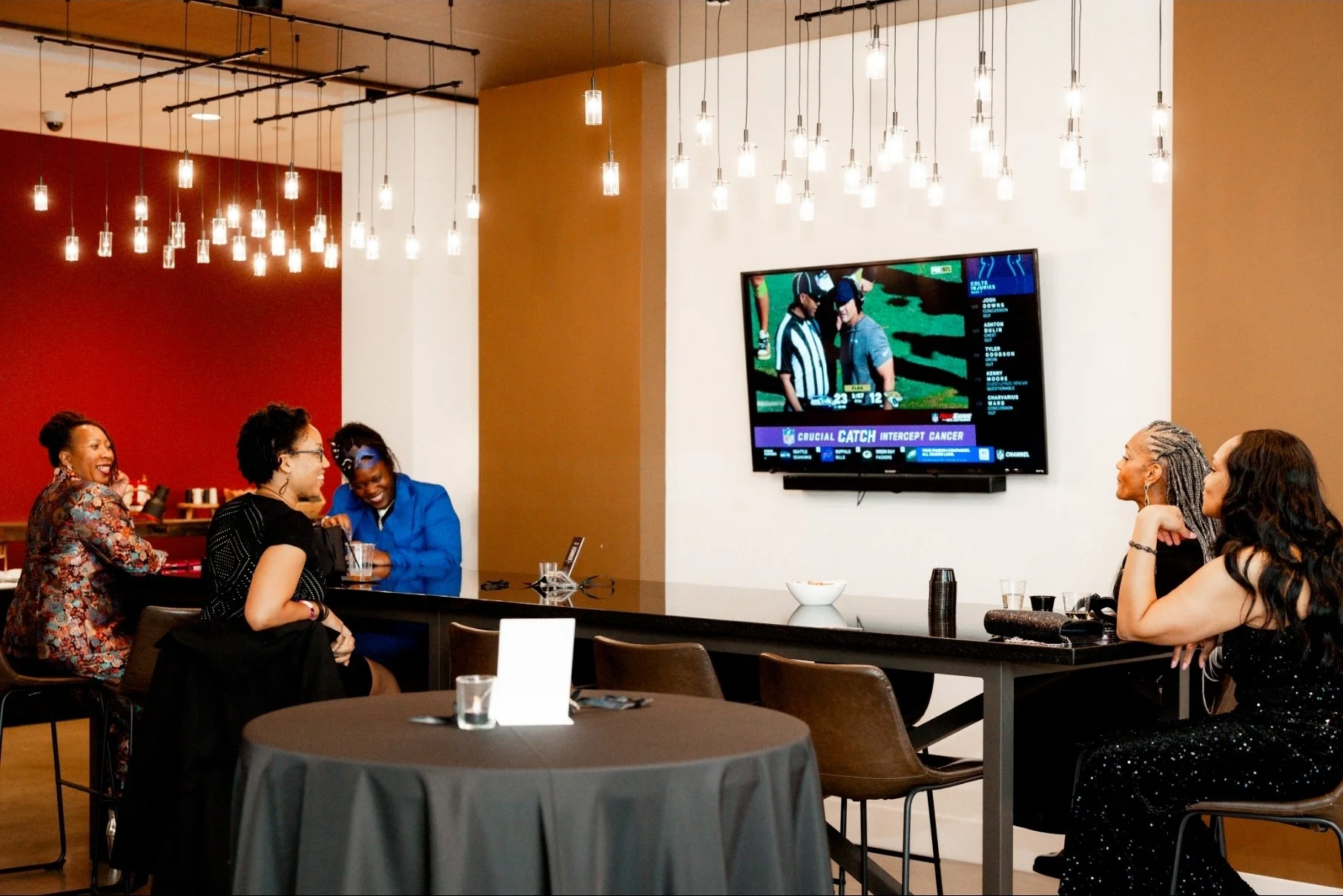 A group of women sitting at a bar in a restaurant or lounge, watching a football game on a wall-mounted TV. The women are smiling and engaged, some in conversation. The interior has modern lighting with hanging pendant lights, and the background feat