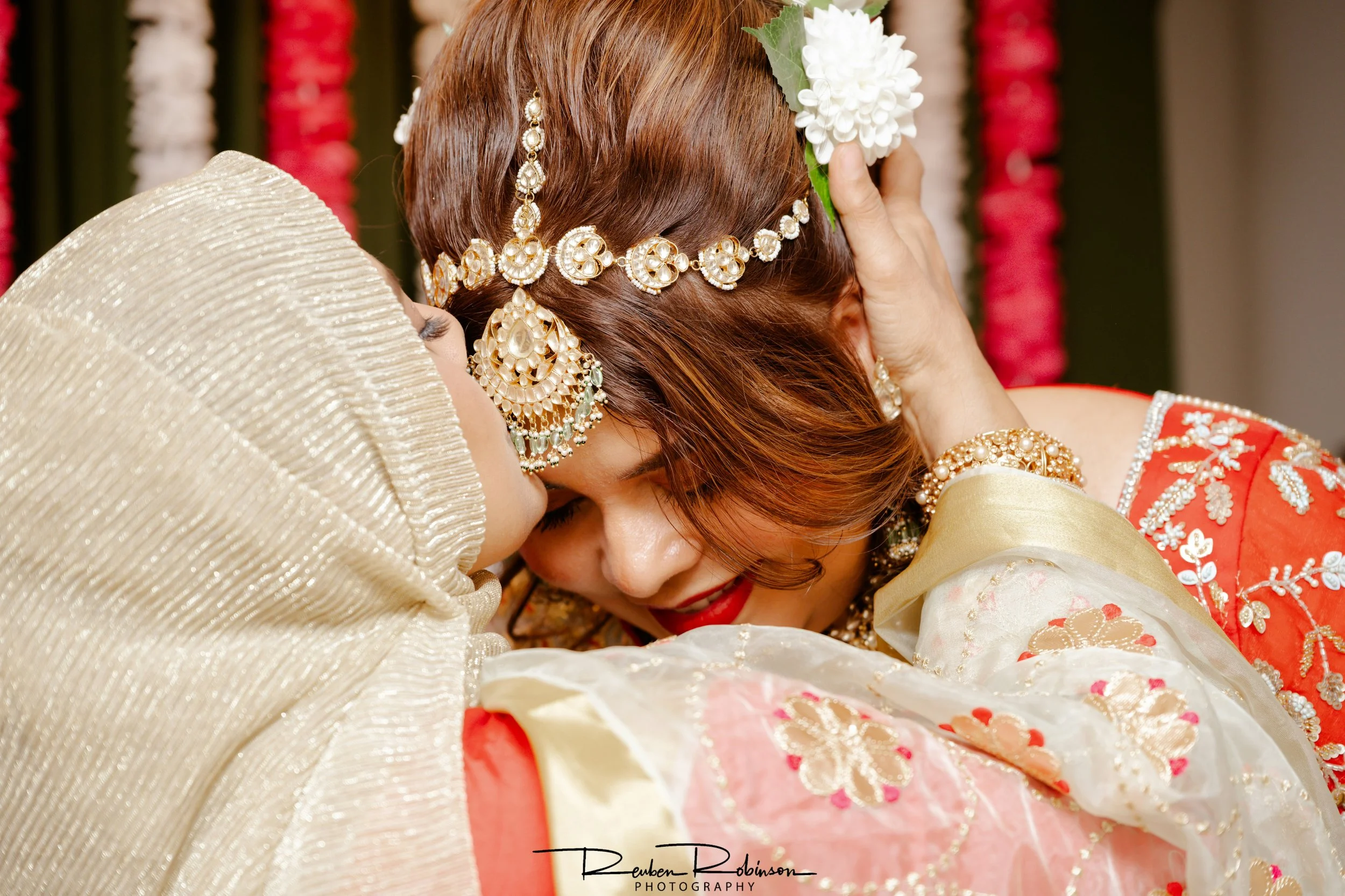 Close-up of an Indian bride and groom in traditional attire, embracing each other during a wedding ceremony. The bride is wearing intricate jewelry, including earrings and a headpiece, and holding a white flower to her head. The groom is dressed in a