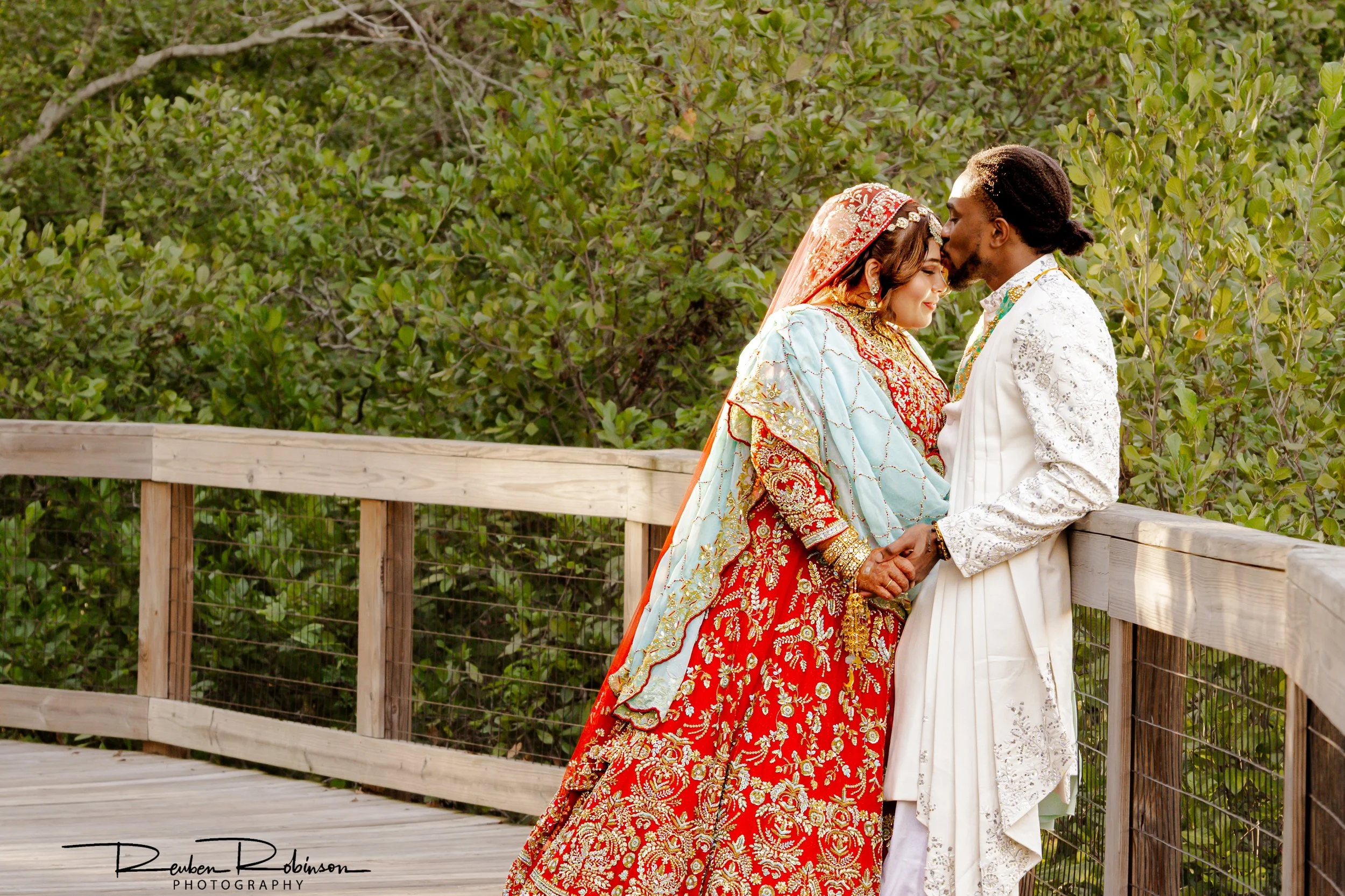 A couple dressed in traditional cultural wedding attire, standing on a wooden bridge, sharing a kiss with their eyes closed. The background features a lush green leafy environment.