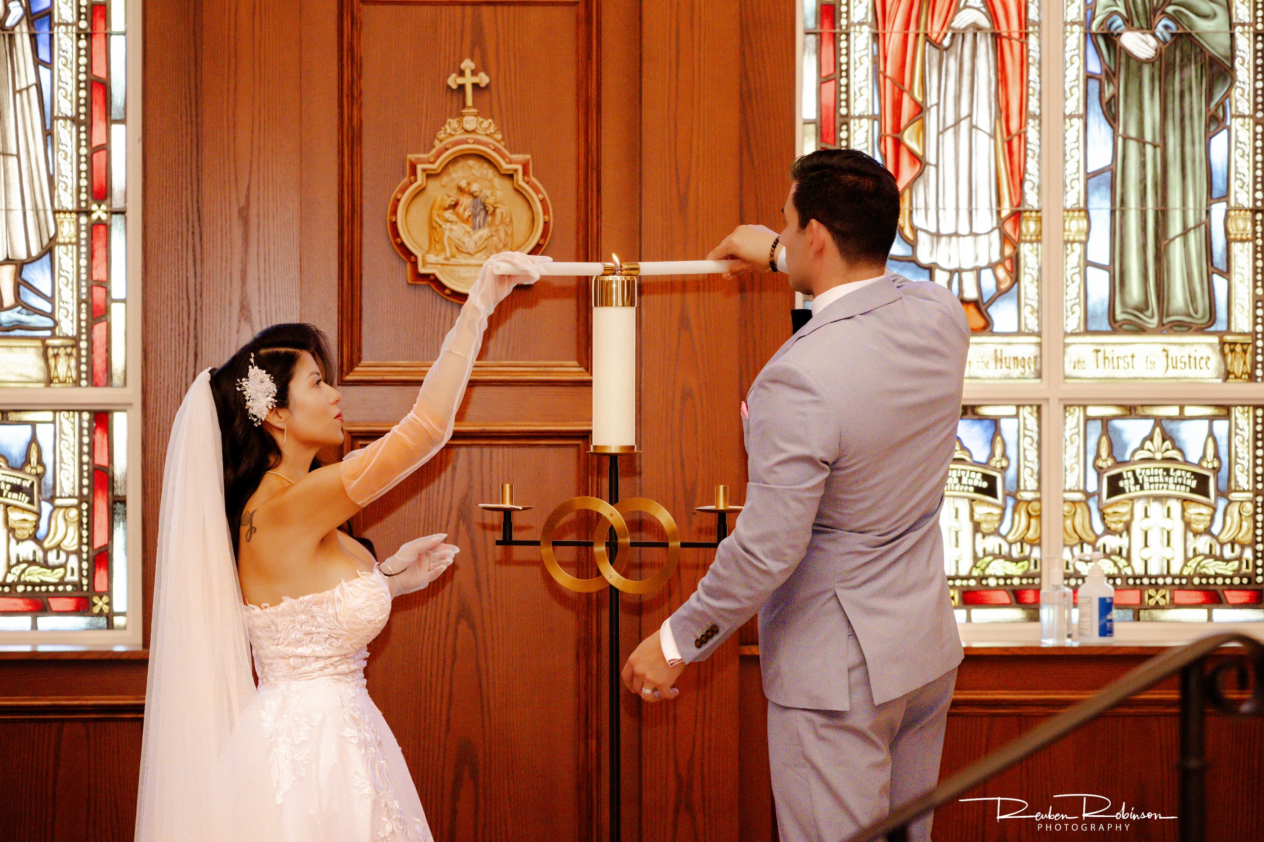 A bride and groom exchange rings at their wedding ceremony inside a church with stained glass windows and wooden wall paneling.