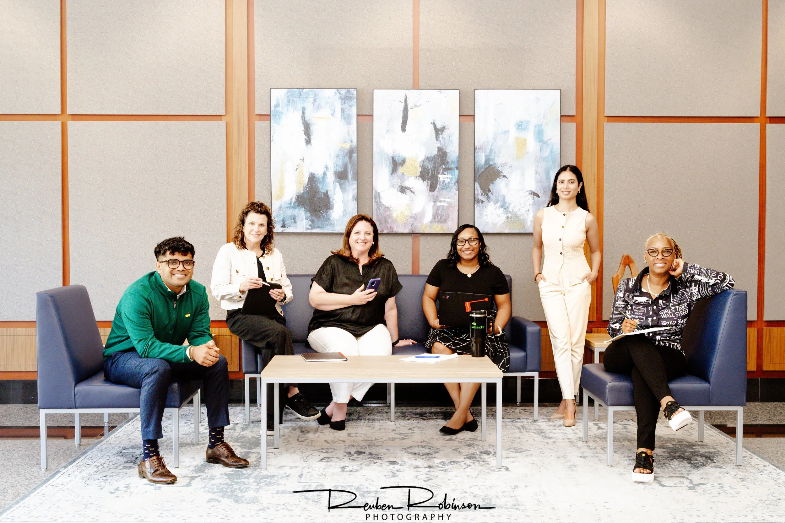 Group of seven diverse women in an office conference room, posing for a photo, with abstract artwork on the wall behind them.