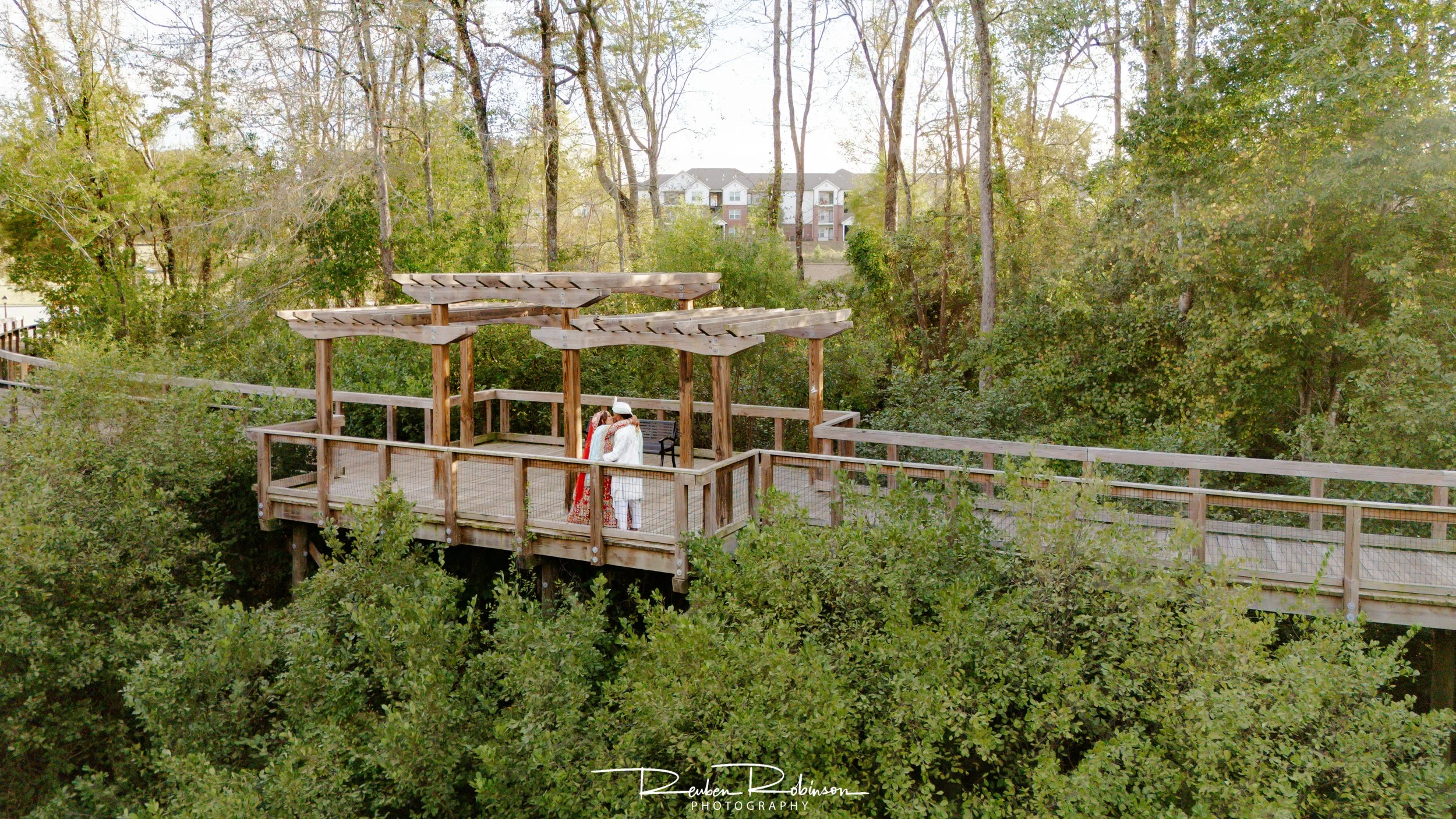 A couple is standing on a wooden deck surrounded by trees, with houses visible in the background.
