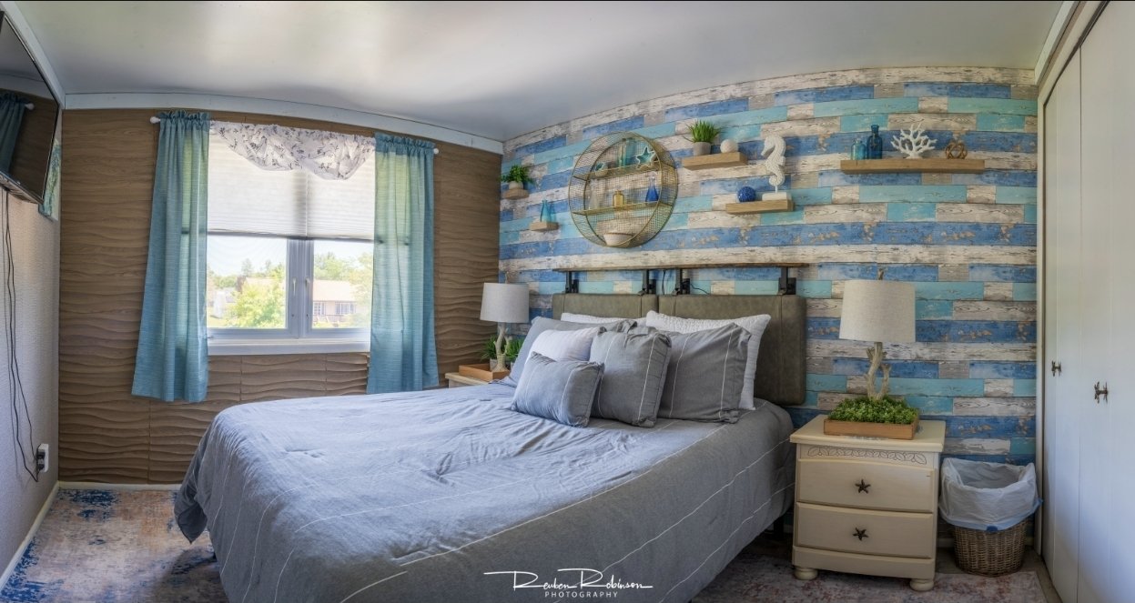 A bedroom with a blue and white rustic accent wall, a large bed with gray bedding and pillows, a nightstand with a lamp and decor, a window with blue curtains, and a closet with sliding doors.