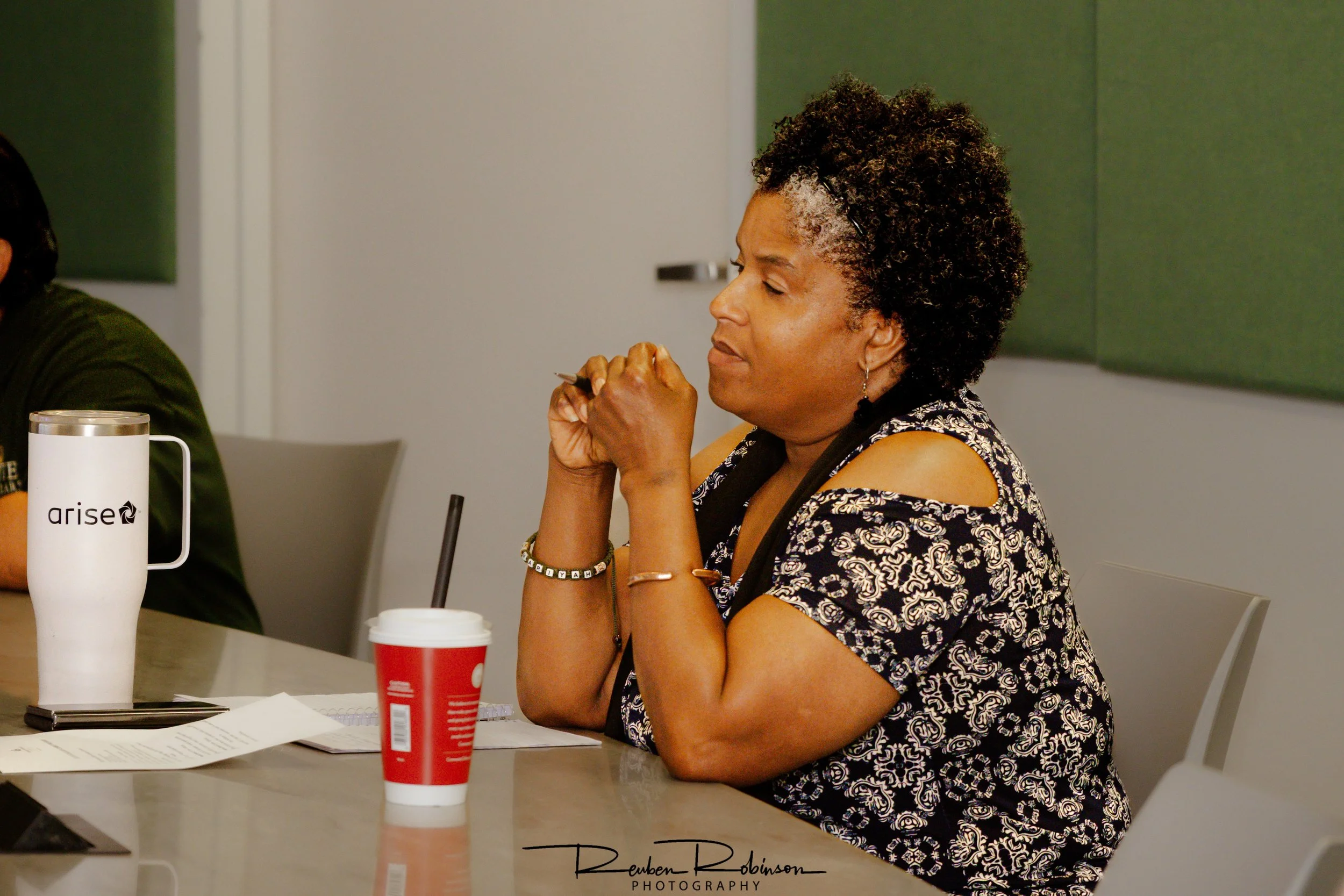 A woman with short, curly hair sitting at a table, with her hands clasped near her face, participating in a discussion or meeting.