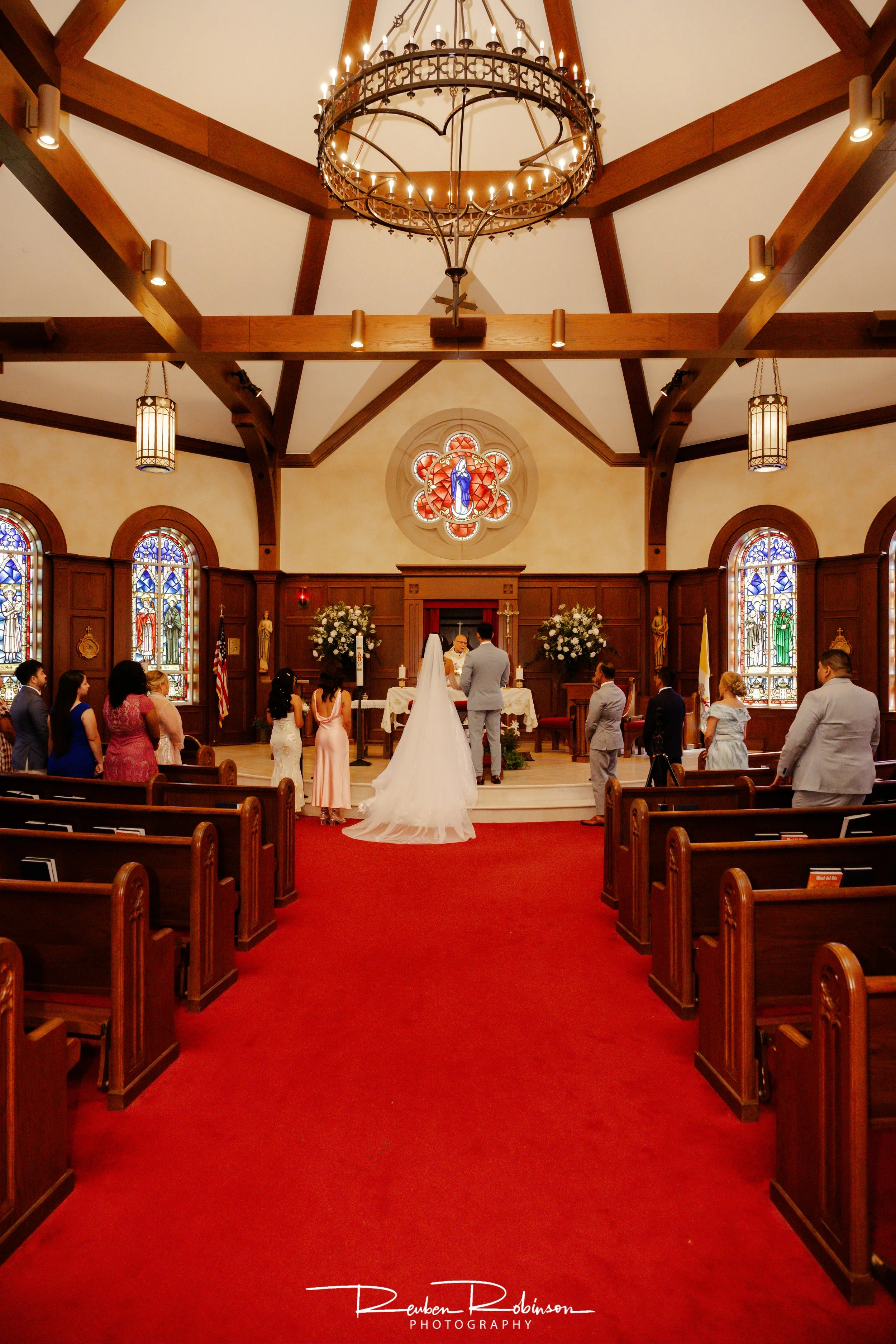 Inside a church decorated for a wedding ceremony, with a bride and groom standing at the altar, surrounded by guests, stained glass windows, and wood-paneled walls.