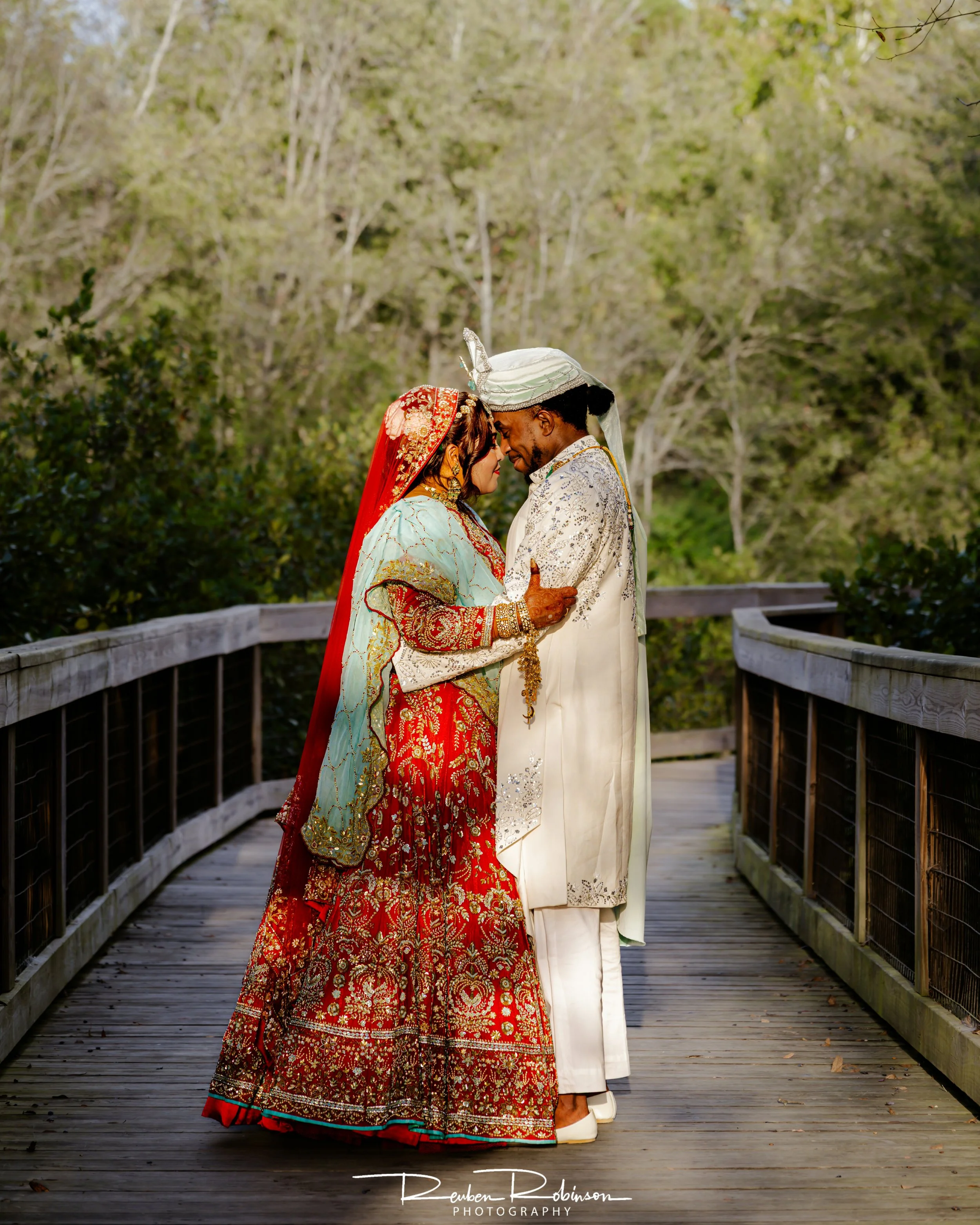 A South Asian couple in wedding attire stands close on a wooden bridge surrounded by trees. The woman is dressed in a red and gold traditional bridal outfit with a red veil, and the man is in a cream-colored sherwani with a white turban. They are tou