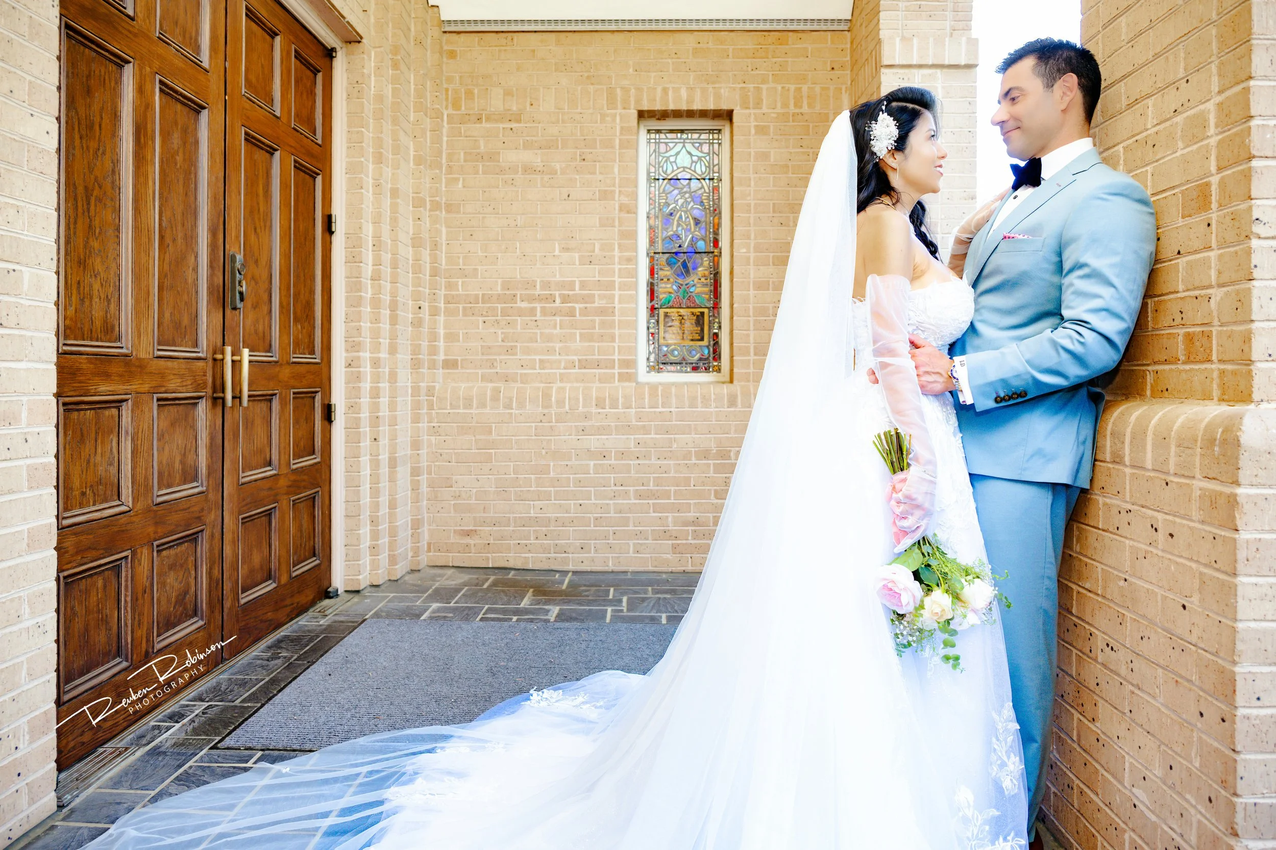 A bride and groom standing close together outside a church, smiling and looking at each other. The bride is wearing a white wedding dress and veil, holding a bouquet of flowers. The groom is in a light blue suit with a bow tie. The scene captures a r