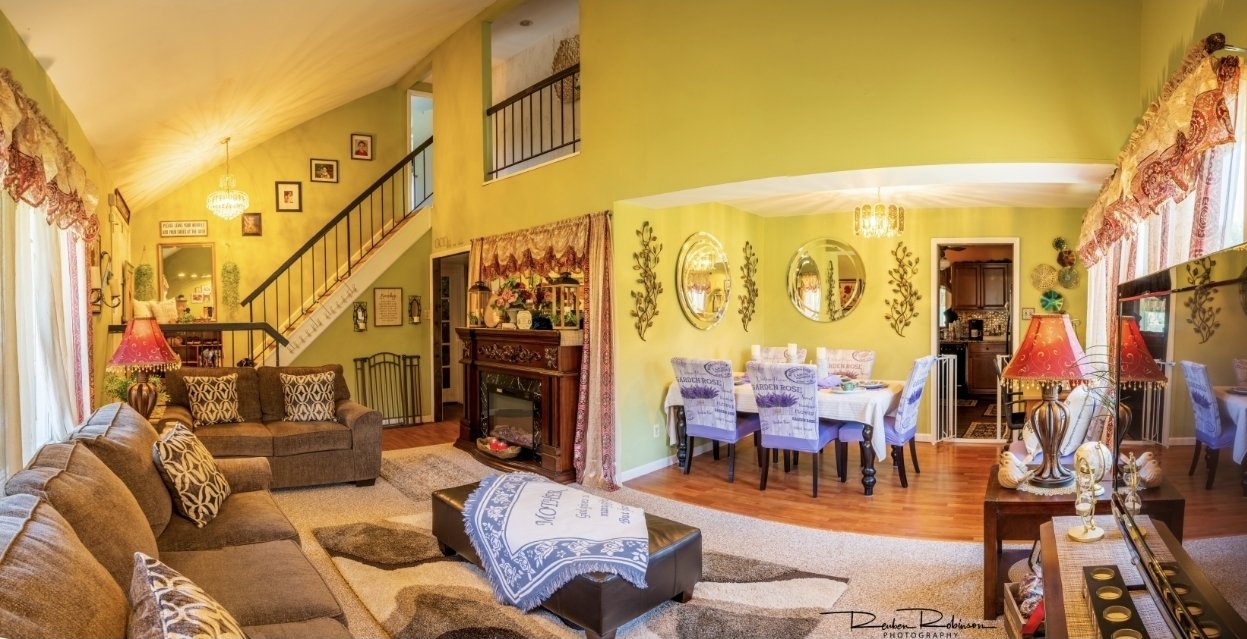 Living room and dining area with yellow walls, brown sofas with patterned pillows, a fireplace, round dining table with eight chairs, and various decorations including lamps, mirrors, and curtains.