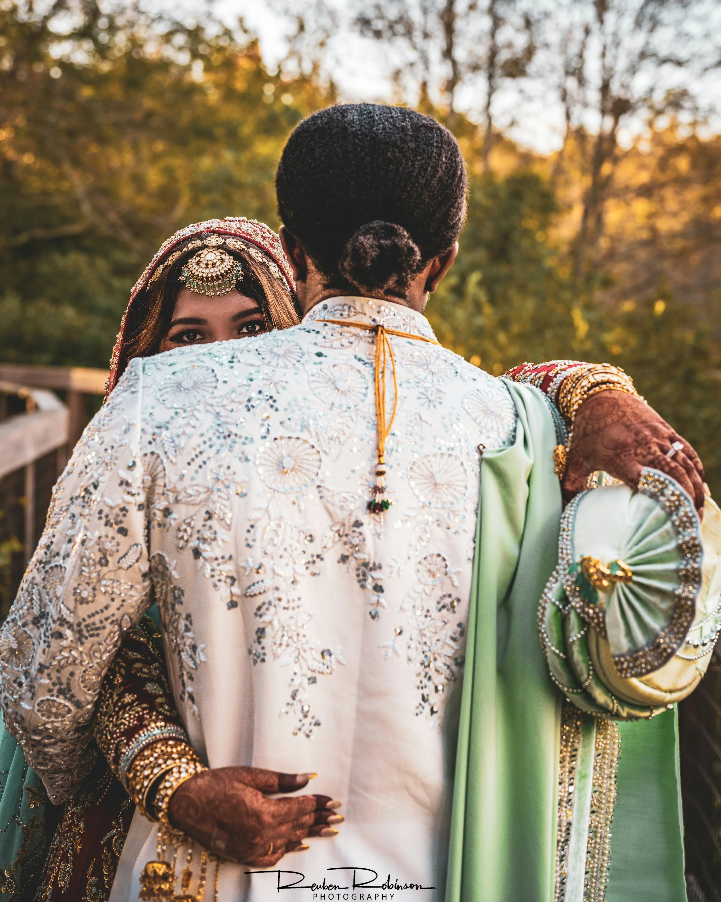 An Indian bride and groom embrace outdoors during sunset, with the bride's face partially visible over the groom's shoulder. The groom wears a white embroidered sherwani, and the bride wears traditional jewelry and henna on her hands.