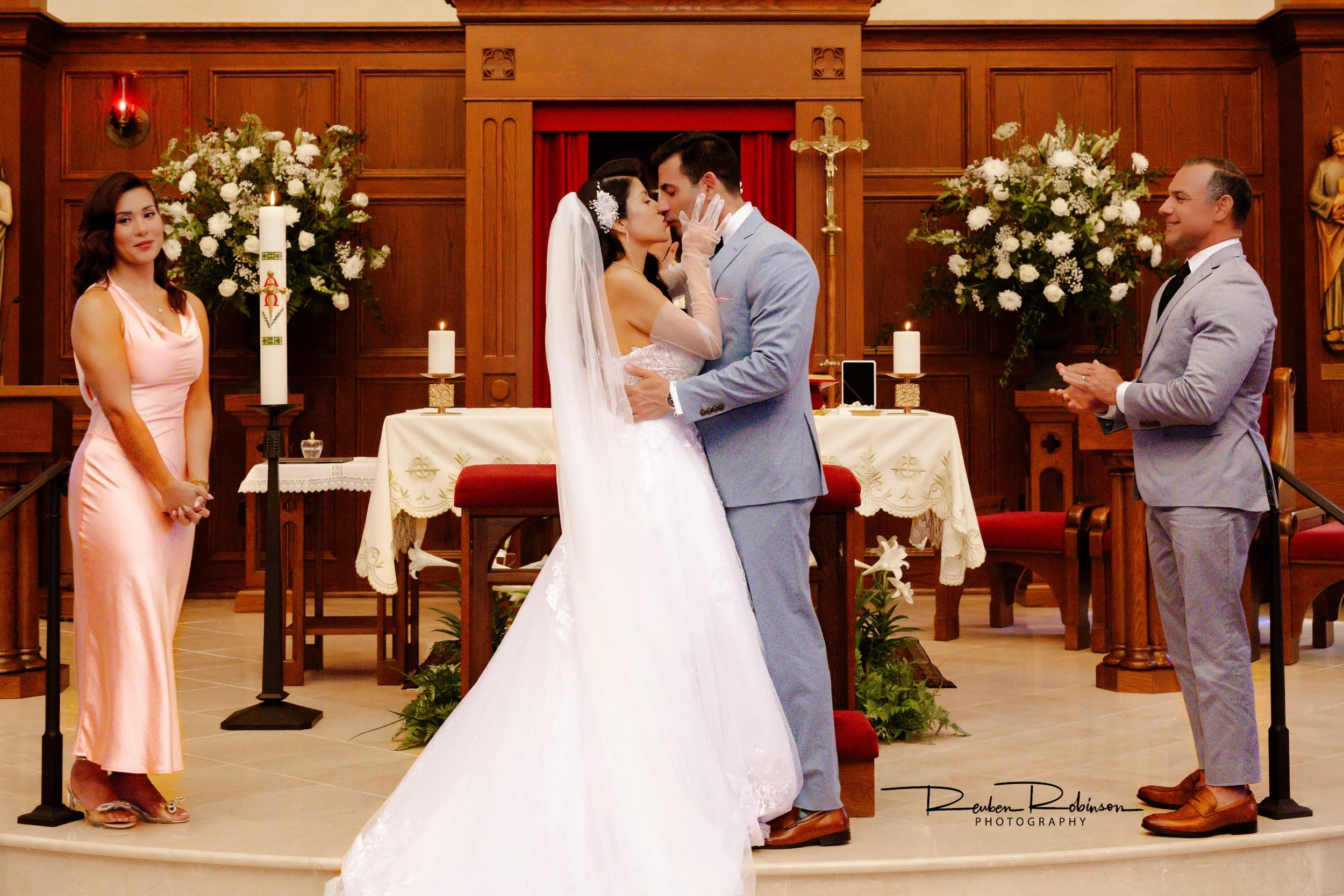 A couple exchanging a kiss at their wedding ceremony in a church with wooden paneling and floral arrangements, with a woman in a pink dress and a man in a gray suit observing.