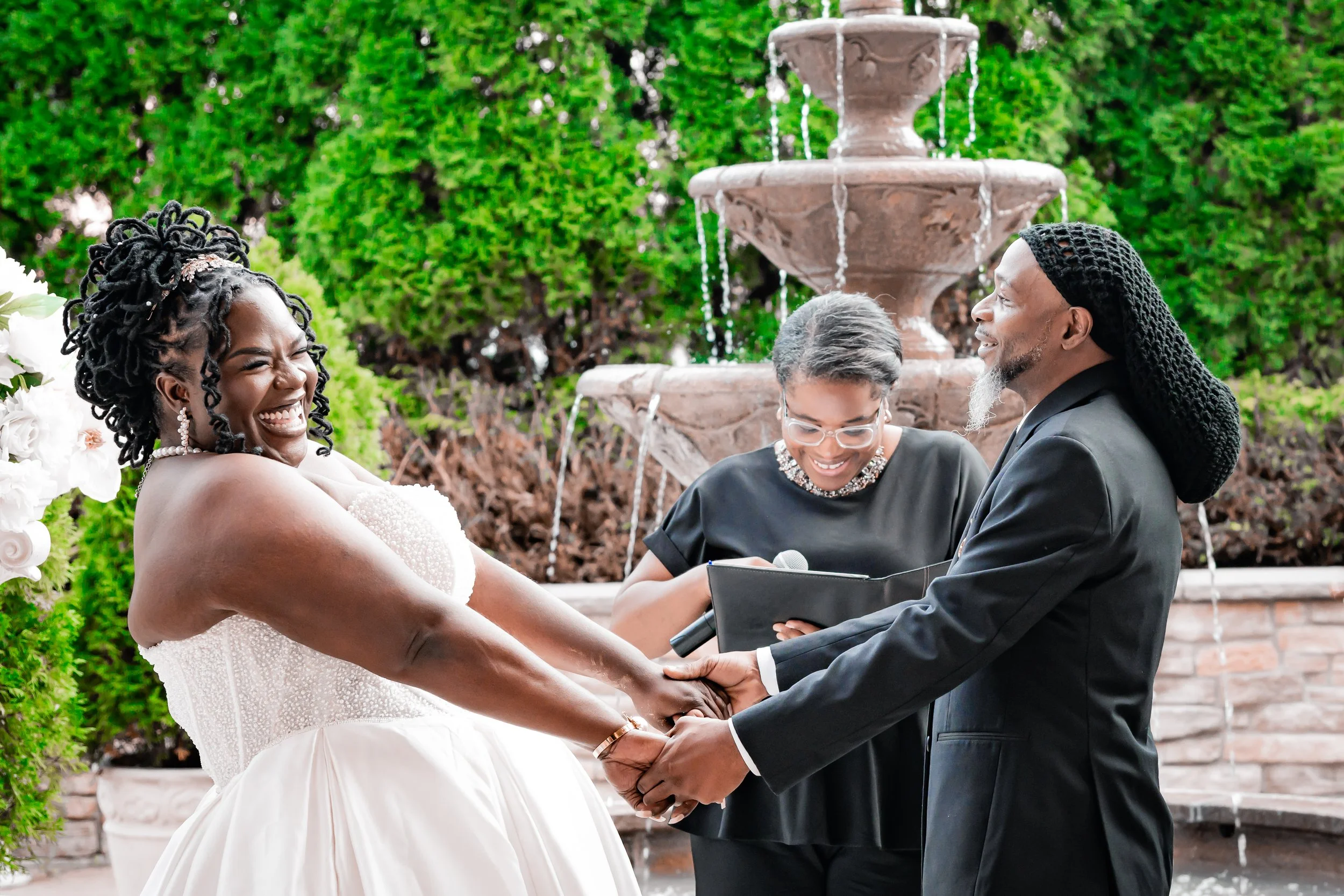 A joyful couple holding hands during their wedding ceremony outdoors with an officiant, and a two-tier fountain in the background.