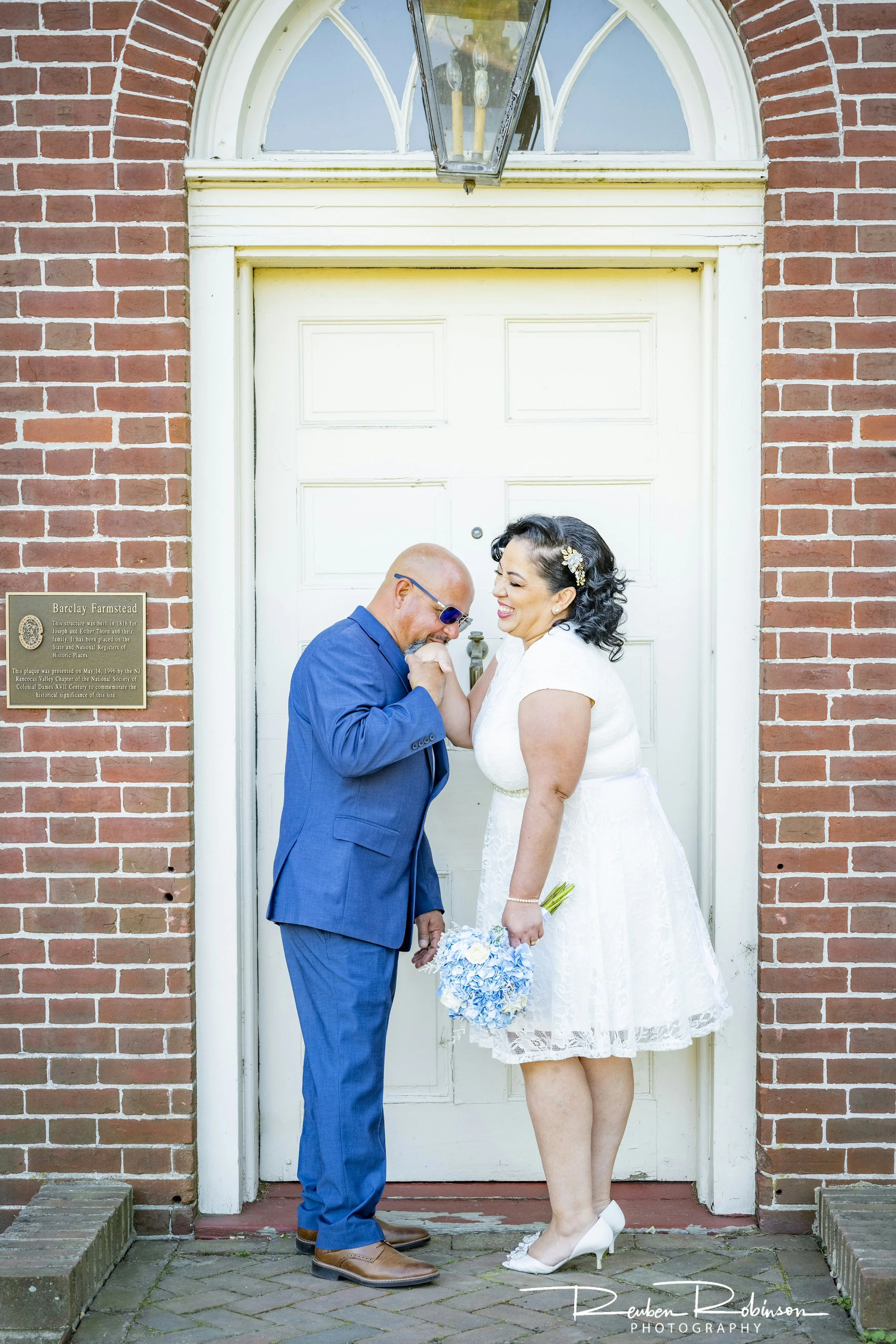 A couple dressed in wedding attire; the bride is holding a bouquet and groom is kissing her hand in front of a brick building.