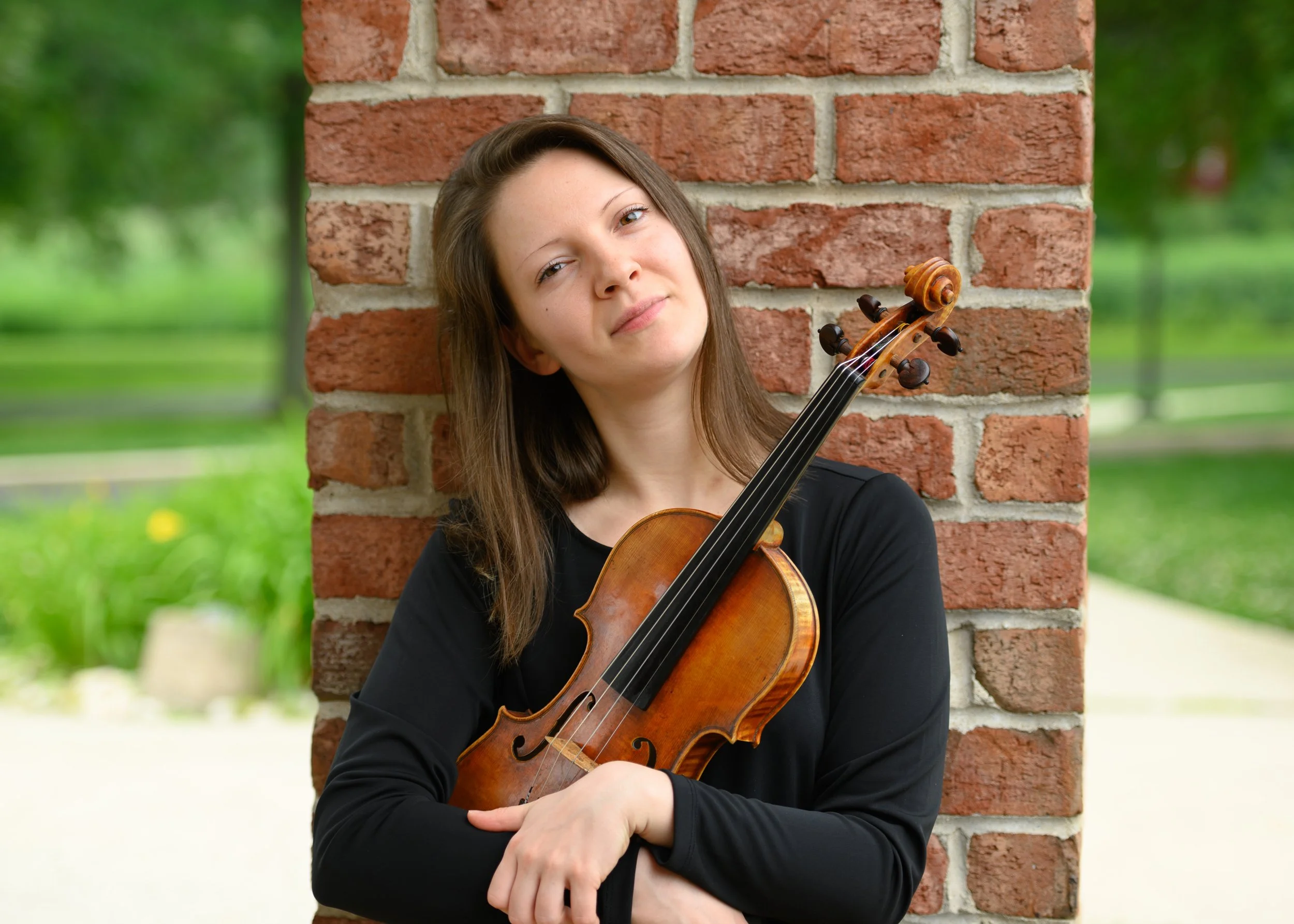 Young woman with long brown hair holding a violin, leaning against a brick wall outdoors with green trees in the background.