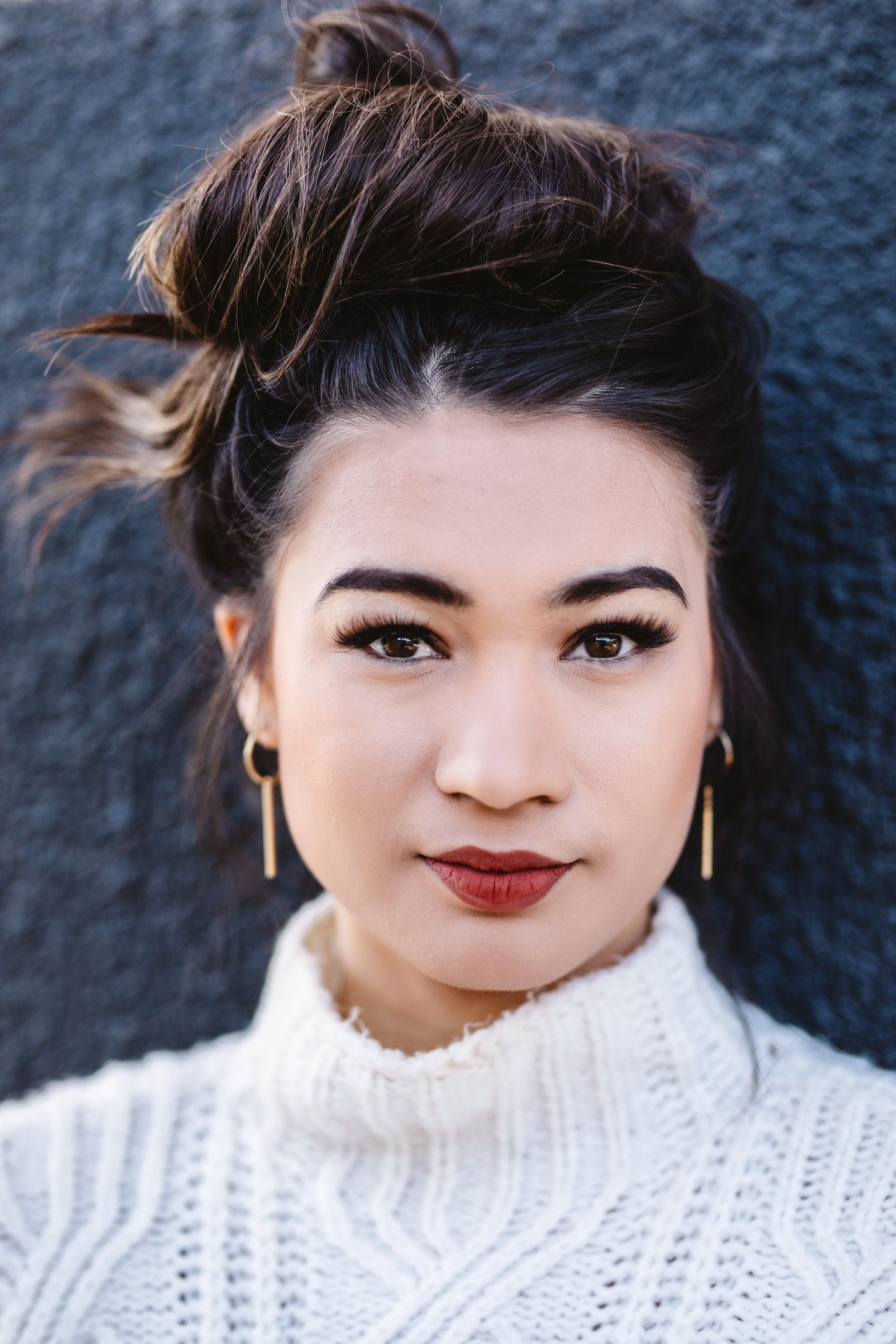 Close-up portrait of a young woman with dark hair styled in a messy updo, wearing a white knit sweater and gold earrings, lying on a dark surface.