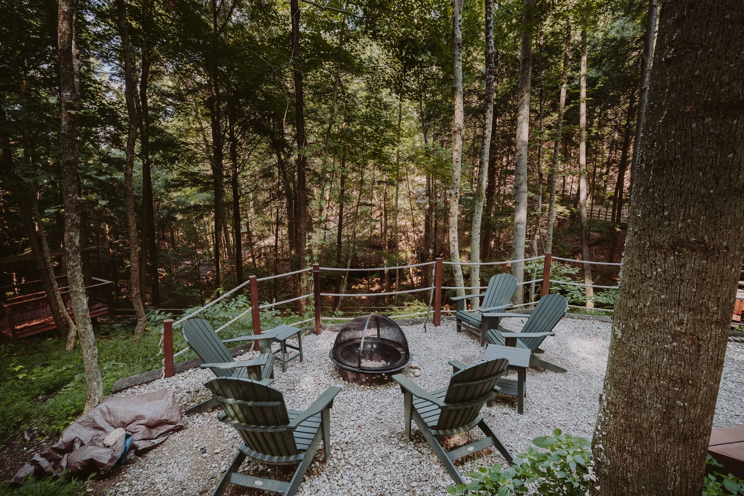 Outdoor seating area with Adirondack chairs arranged around a fire pit on gravel, surrounded by tall trees in a forest.