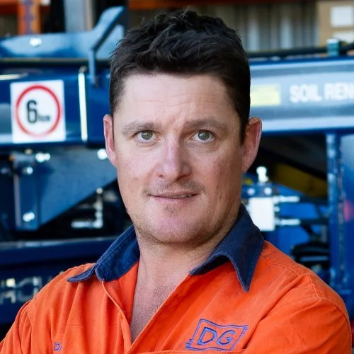 A man in an orange work shirt standing outdoors in front of machinery.