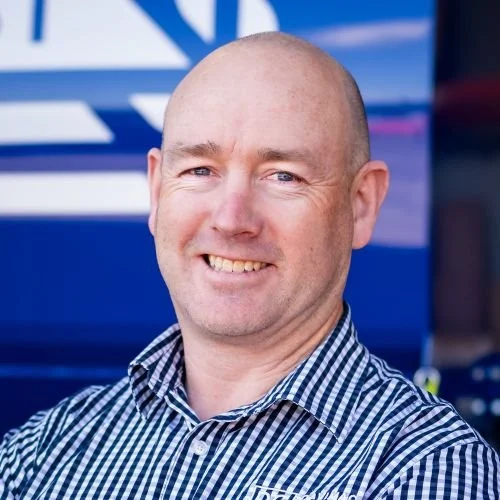 A man short hair smiling, wearing a blue and white checkered shirt, standing outdoors with a blurred industrial or mechanical background.