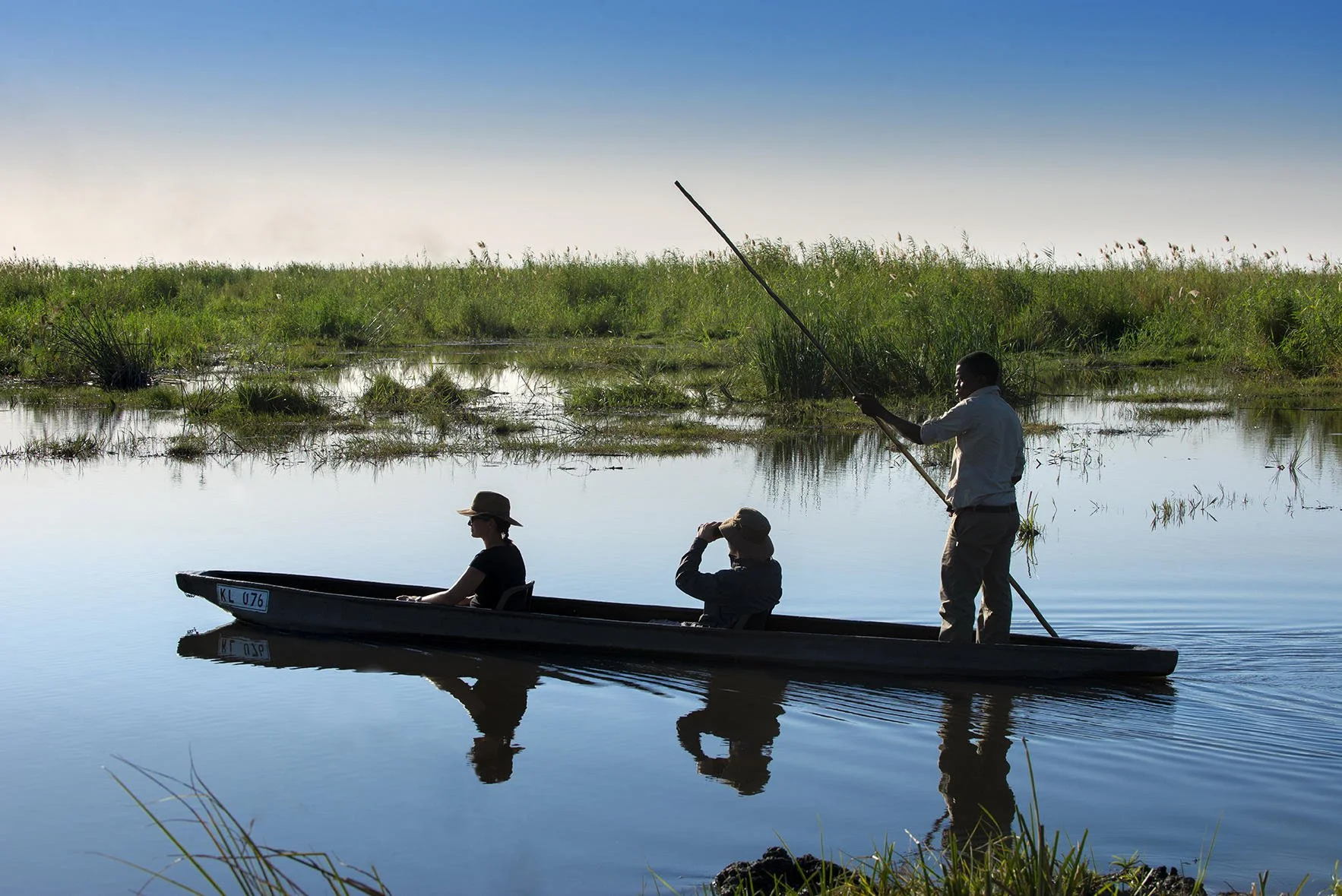 Three people in a boat on calm water, with one person standing and paddling with a pole, and two sitting, one with a hat and glasses, the other with a hat, against a backdrop of greenery and a clear blue sky.