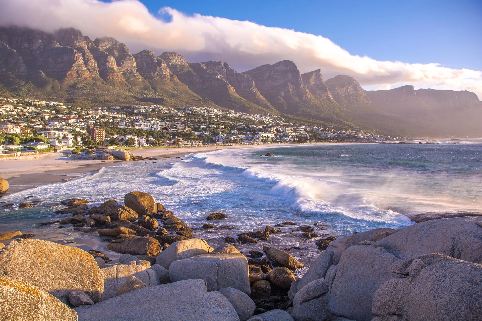 Scenic view of a beach with large rocks in the foreground, waves crashing on the shore, a town along the coast, and mountains covered in clouds in the background.