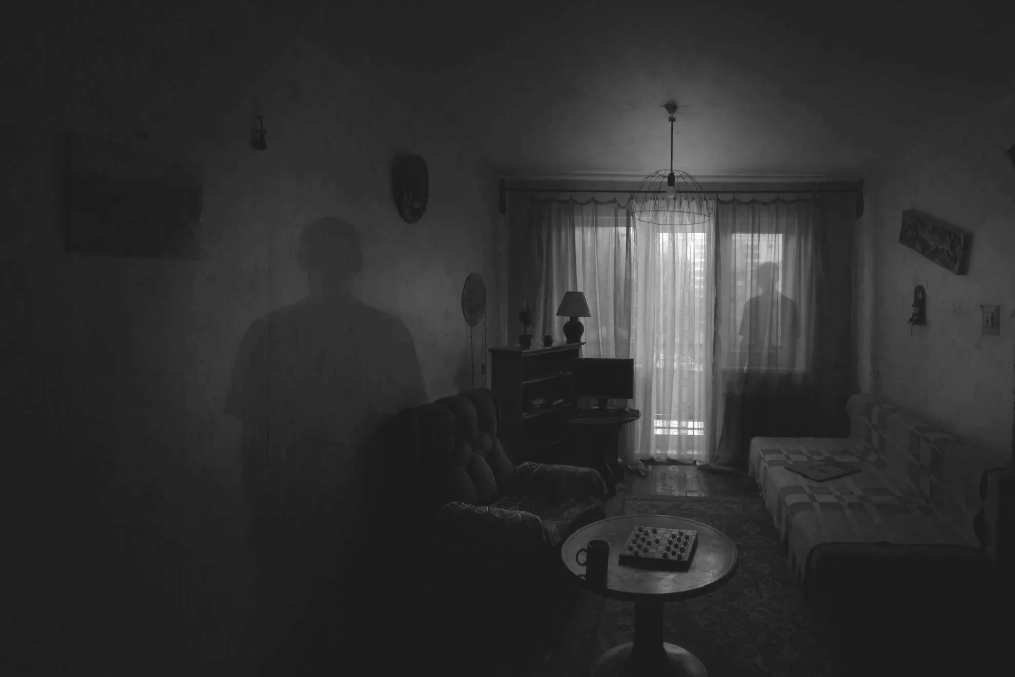 Sala de estar com cortinas, sofá, mesa com jogo de tabuleiro, televisão e uma luminária, com duas sombras de pessoas visíveis através da janela.