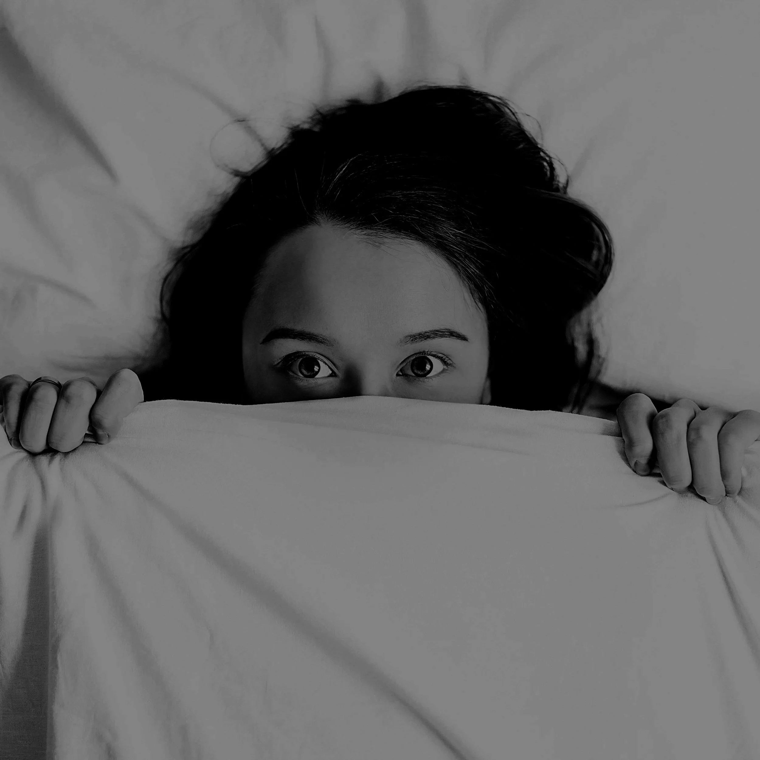 Pessoa de cabelo escuro segurando a cama, com olhos abertos, observando o observador em uma imagem em preto e branco.