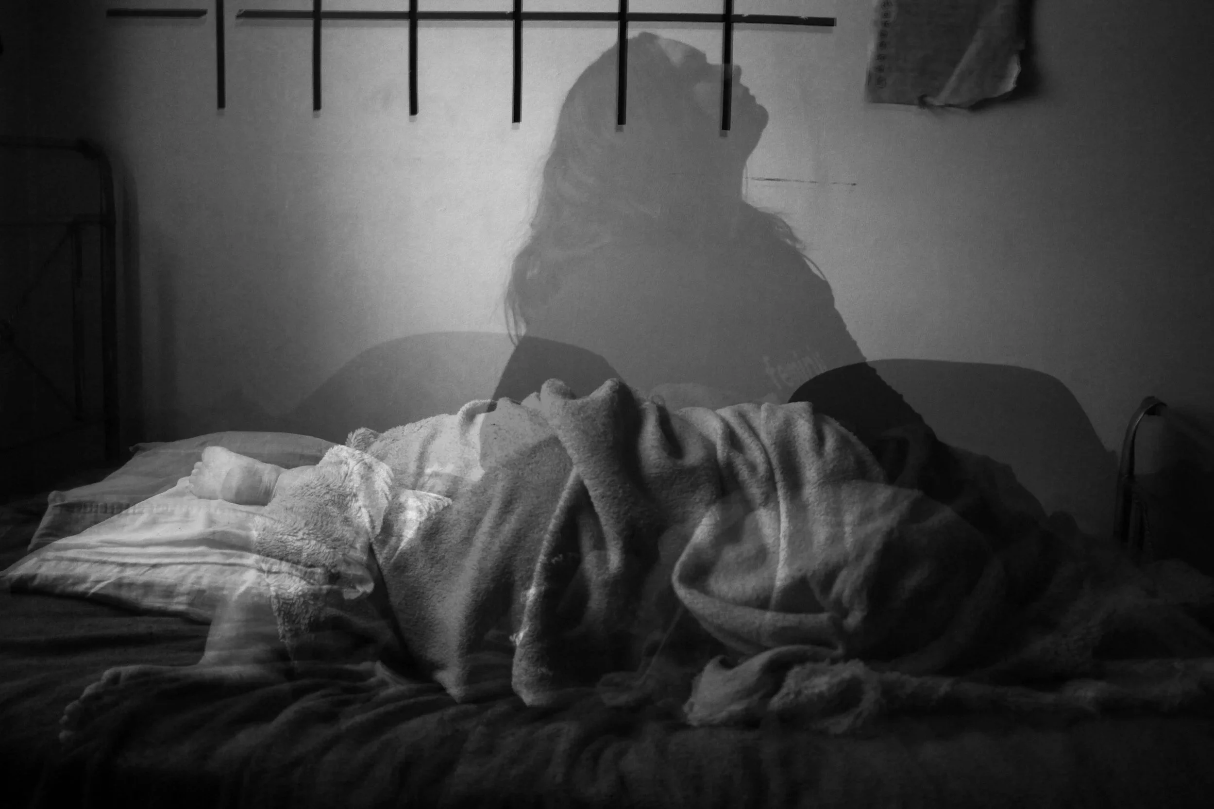 Imagem em preto e branco de uma pessoa deitada em uma cama com uma manta, com uma sombra projetada na parede atrás dela, criando uma silhueta de uma pessoa de pé.