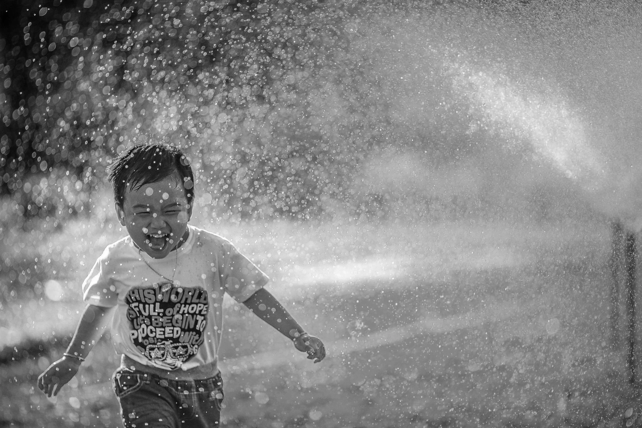 Criança brincando na água, sorrindo e se divertindo, com gotas de água ao redor em cenário ao ar livre.