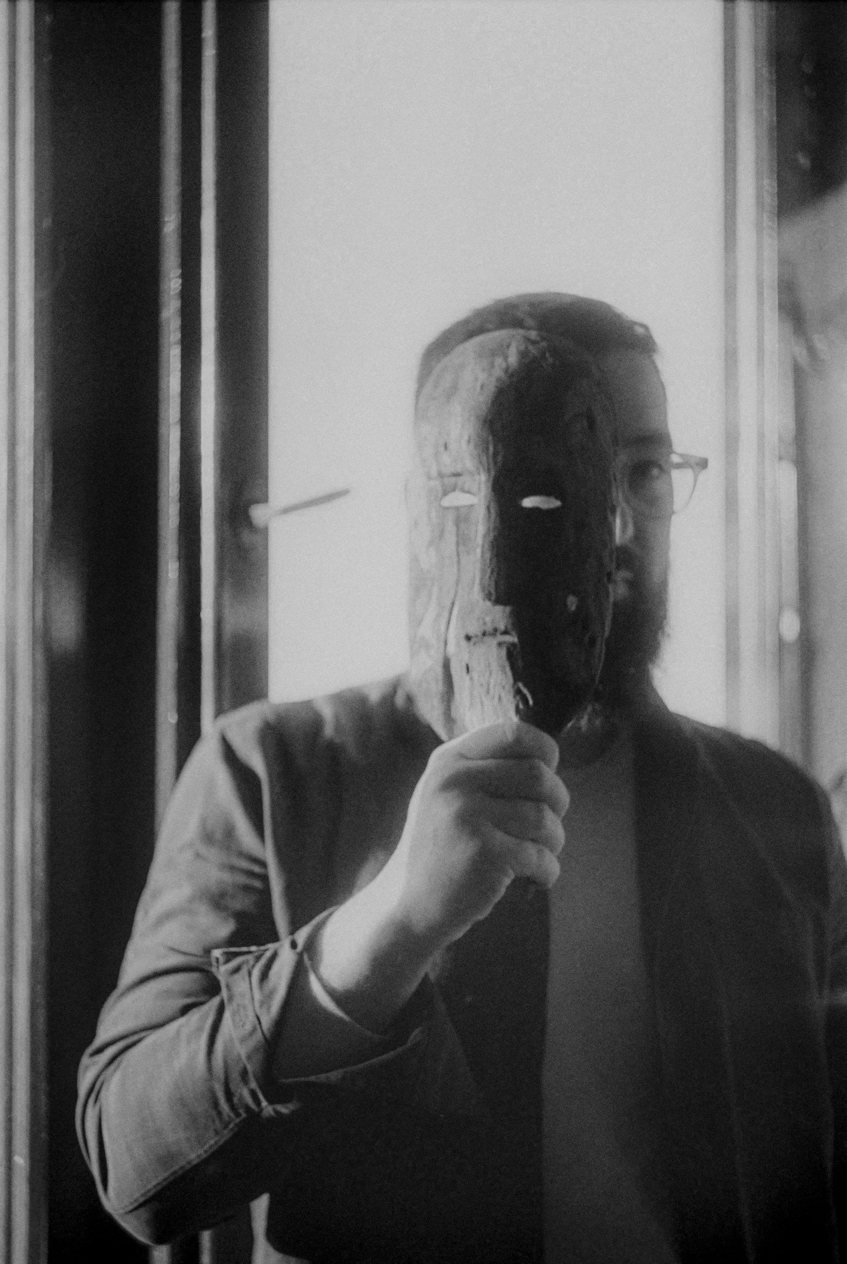 Homem com barba e óculos segurando uma máscara de madeira na frente do rosto, no reflexo de um espelho