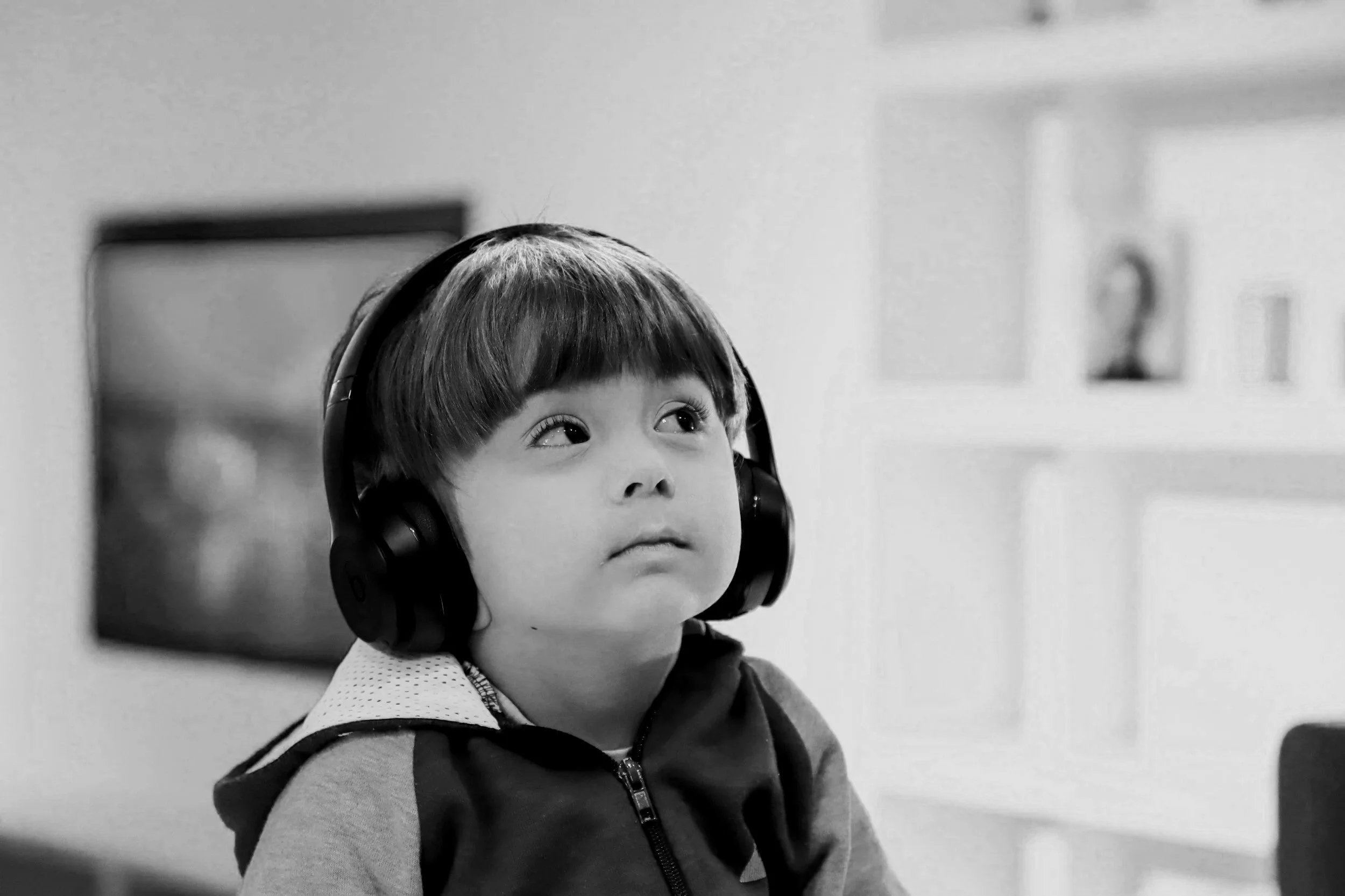 Menino com cabelo curto usando fones de ouvido pretos e uma jaqueta cinza escura, sentado em um ambiente interno, expressão neutra, olhando para o lado.