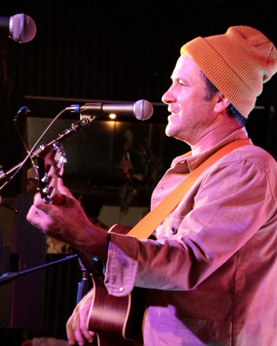 Man wearing a yellow beanie playing guitar and singing into a microphone on stage.