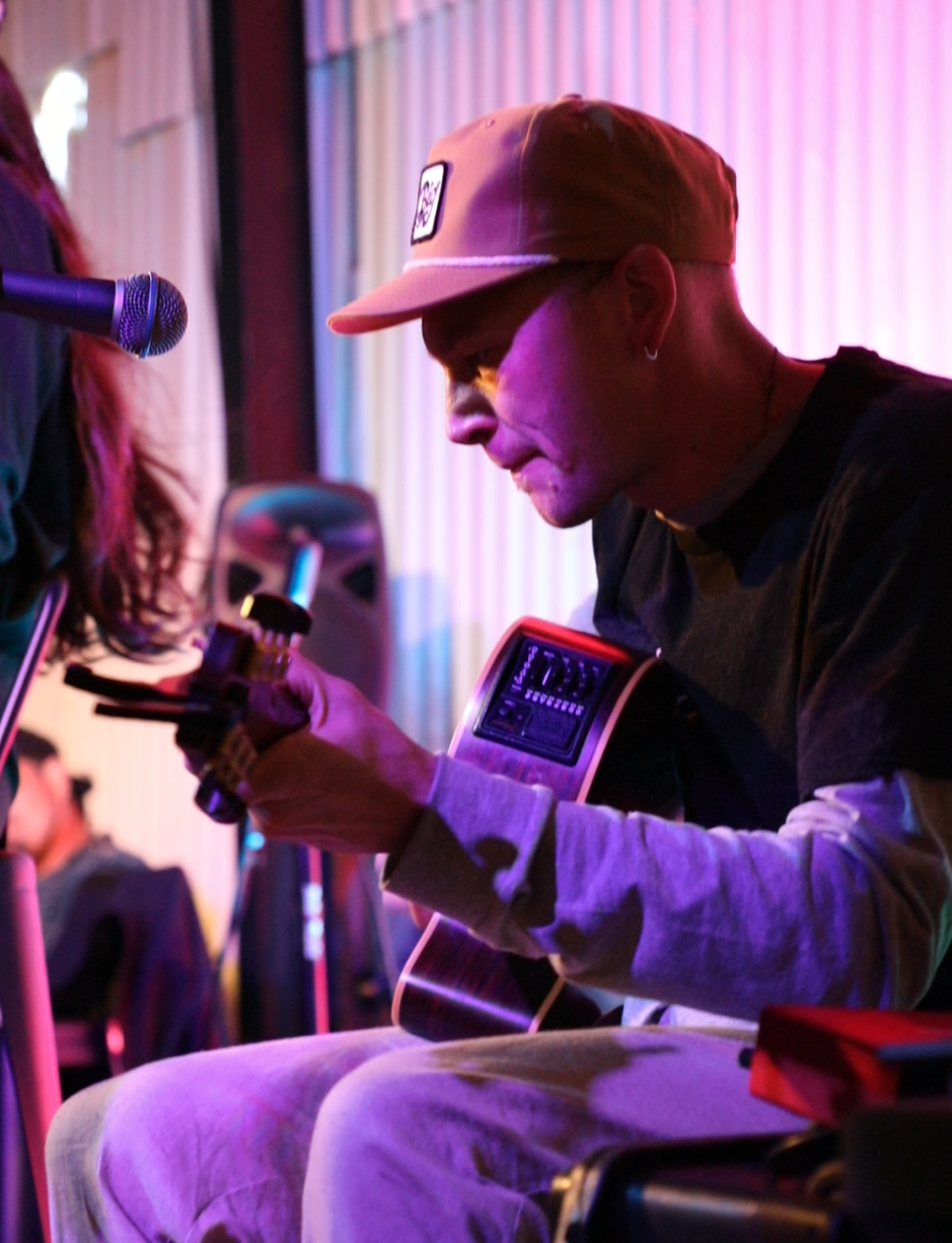 A person wearing a beige cap and earrings is sitting and playing an acoustic guitar with a capo on the fretboard. They are in a room with colorful, semicircular, vertically striped curtains illuminated by pink, purple, and blue lighting. A person wit