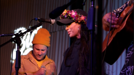 A woman wearing a floral crown and black jacket standing on stage with two men, one in a yellow beanie, and another playing a guitar, in a performance setting