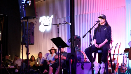 A woman in dark clothing and sunglasses sitting on a chair on stage with a microphone, surrounded by musical equipment and a seated audience watching her perform.