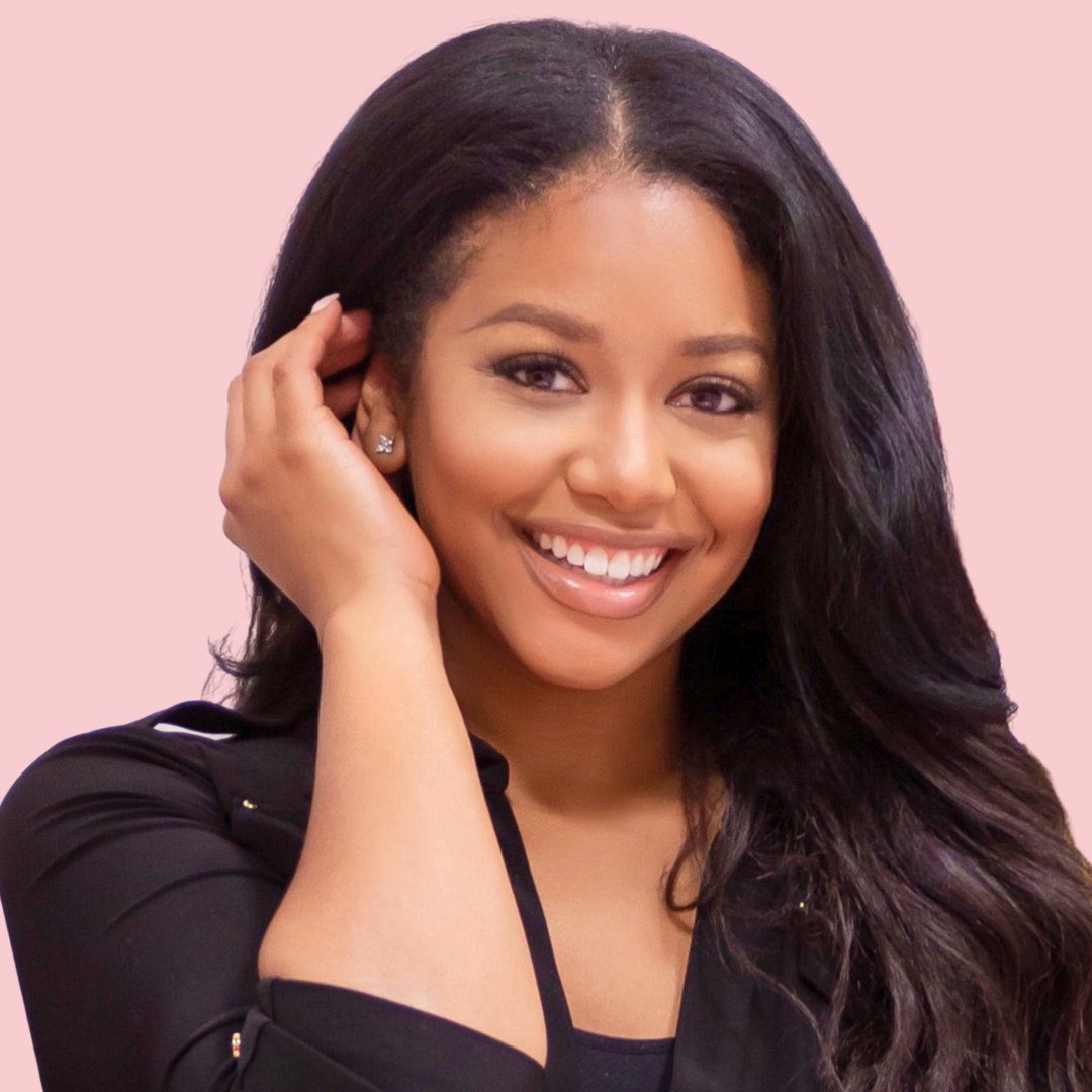 A smiling woman with long dark hair, wearing a black top, adjusting her earring, against a light pink background.