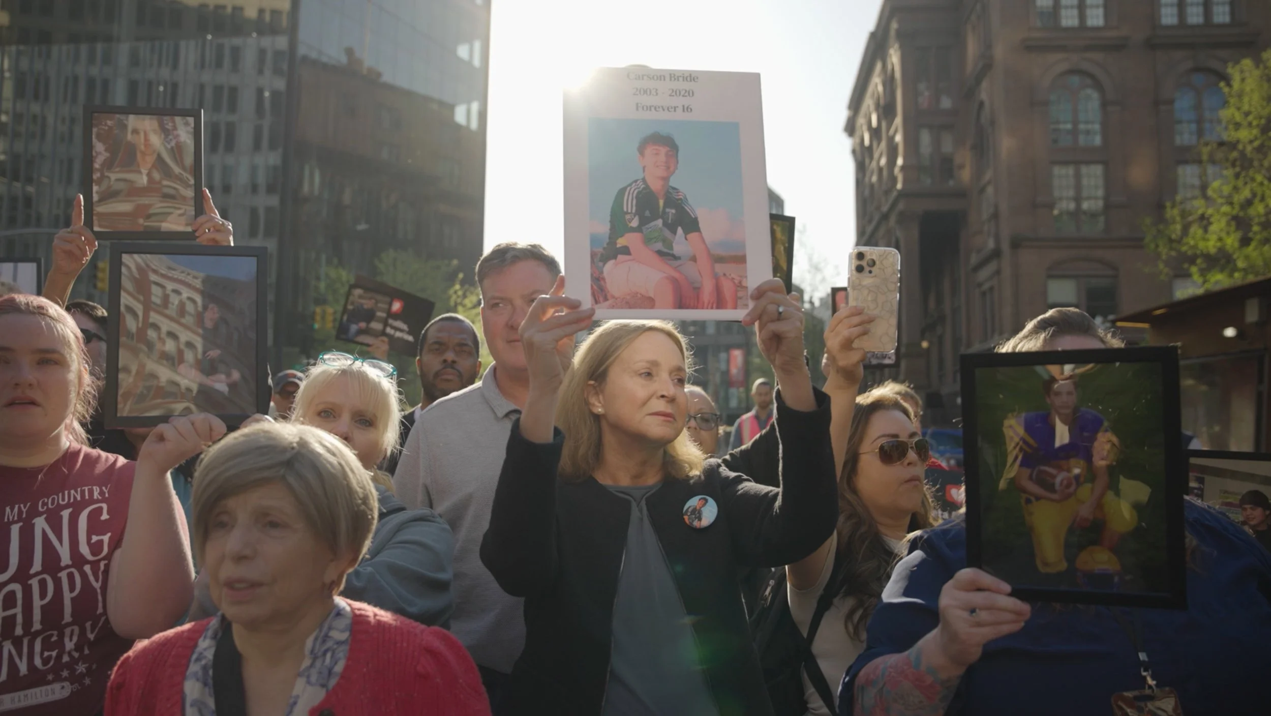 A crowd of people at a memorial march, some holding framed photographs of loved ones, with one person holding a sign with a photo of a young person and the text 'Carson Bride 2003-2020 Forever 16'.