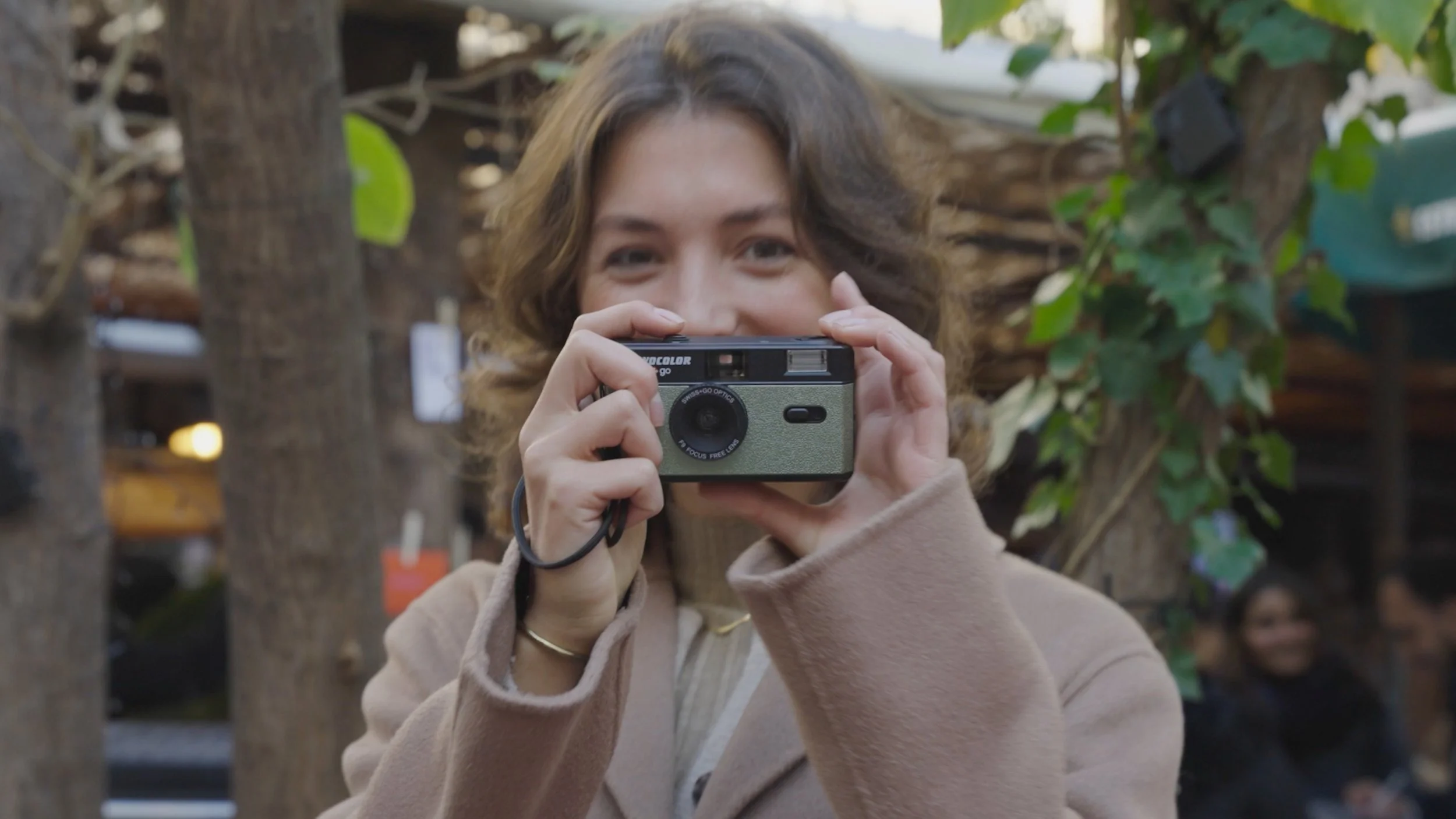 A young woman with curly brown hair pointing a vintage film camera toward the viewer, outdoors surrounded by green leaves and brown tree trunks, smiling with her eyes visible above the camera.