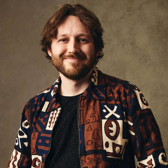 A man with brown hair and beard smiling, wearing a patterned jacket over a black speckled shirt, standing against a neutral background.