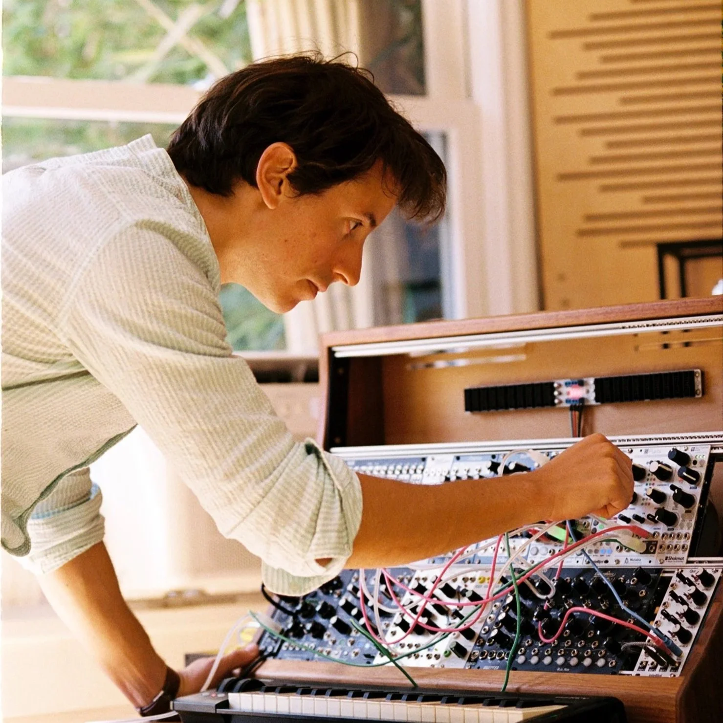 A person with brown hair adjusting a modular synthesizer with many knobs, cables, and a keyboard in front of a window with greenery outside.