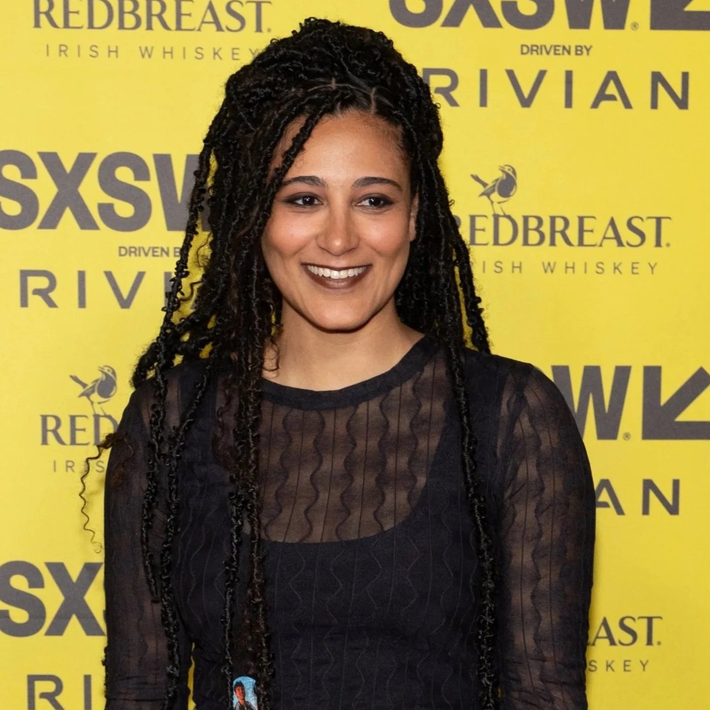 A woman with long twisted braids, smiling in front of a yellow backdrop with logos and text for SXSW, RedBreast Irish Whiskey, and Driven by Rivian.