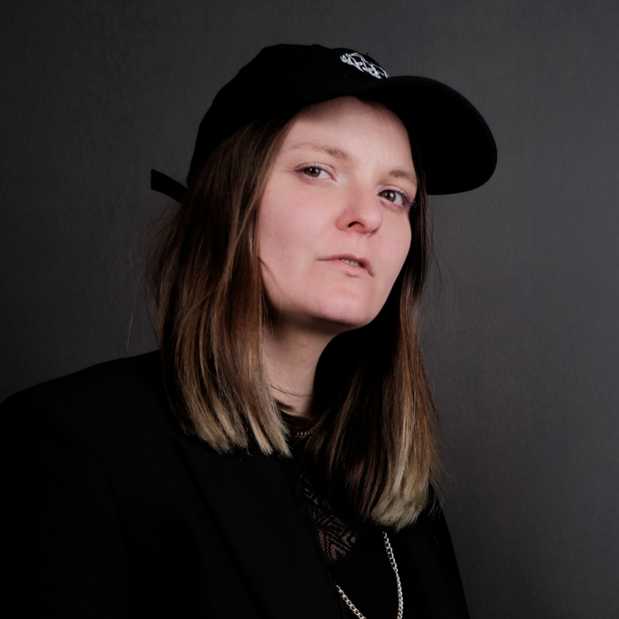 A young woman with long brown hair wearing a black cap and black clothing against a dark background