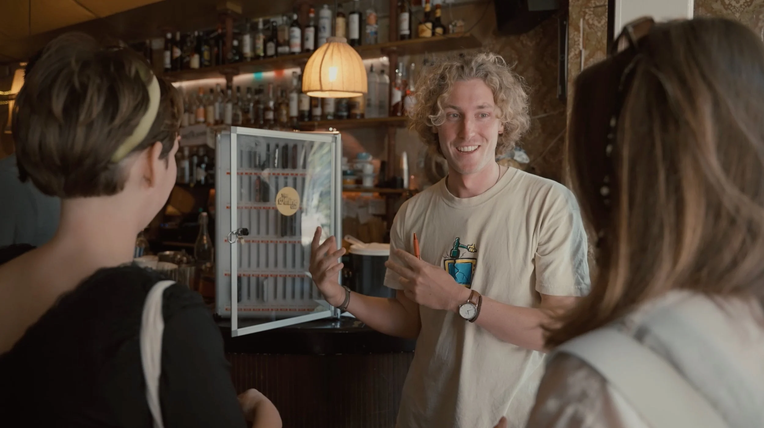 A man with curly blonde hair is talking to two women inside a bar or cafe. The man is smiling and gesturing with his hands, and there are shelves with bottles behind him.