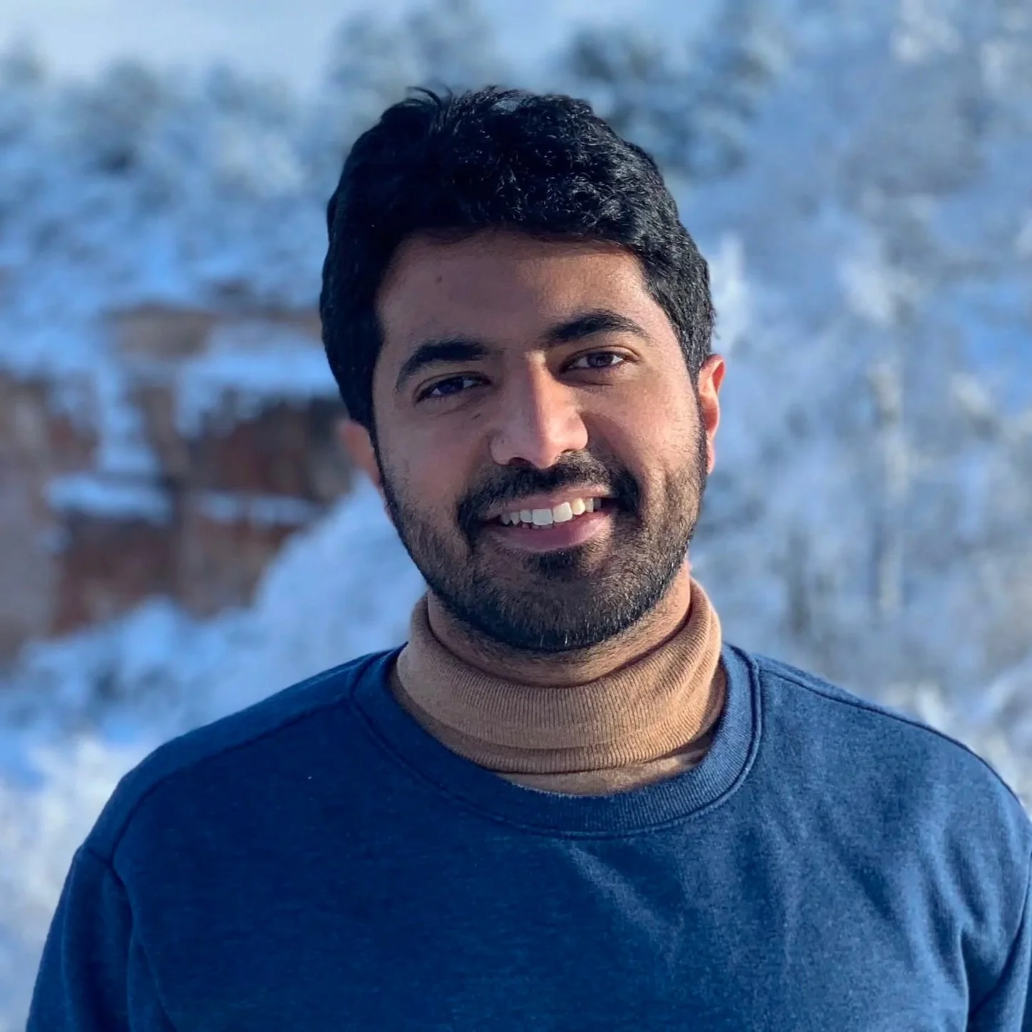 A man with dark hair and a beard smiling outdoors with waves in the background.